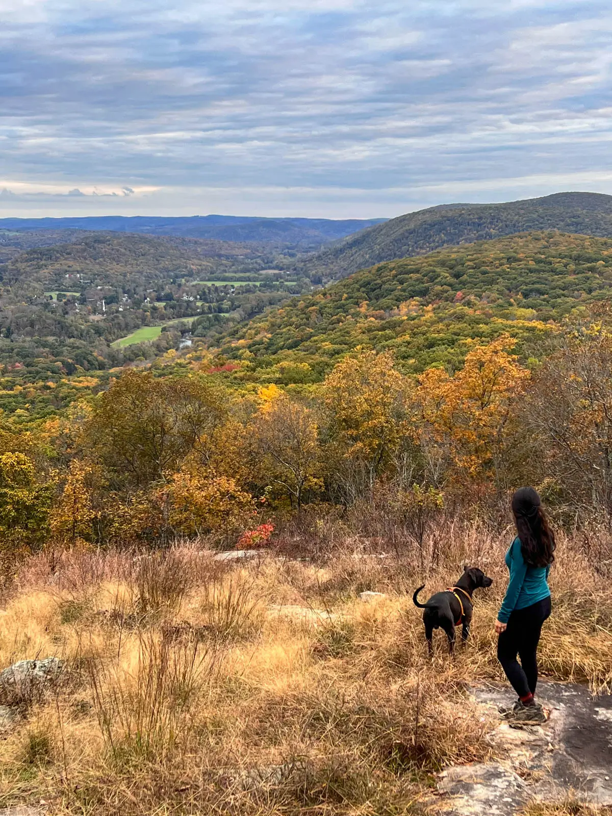 woman in long-sleeve dark green top on mountaintop view from hike on appalachian trail in connecticut in fall with green and orange colored trees and leaves in the distance and cloudy skies above.