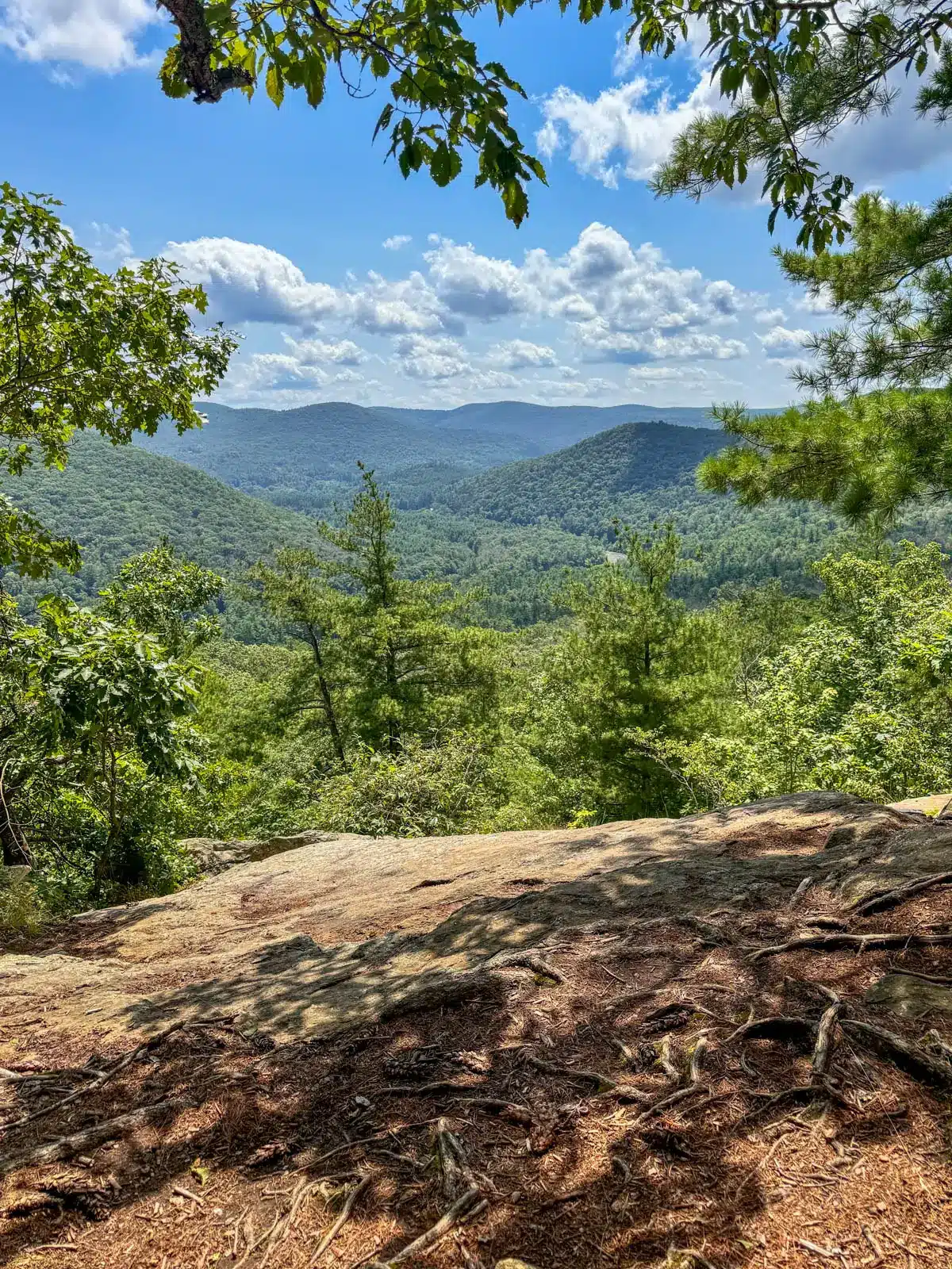view from mountaintop in summer on appalachian trail in connecticut looking out and rolling green hilltops with blue skies and fluffy clouds above and green leaves framing the photo.