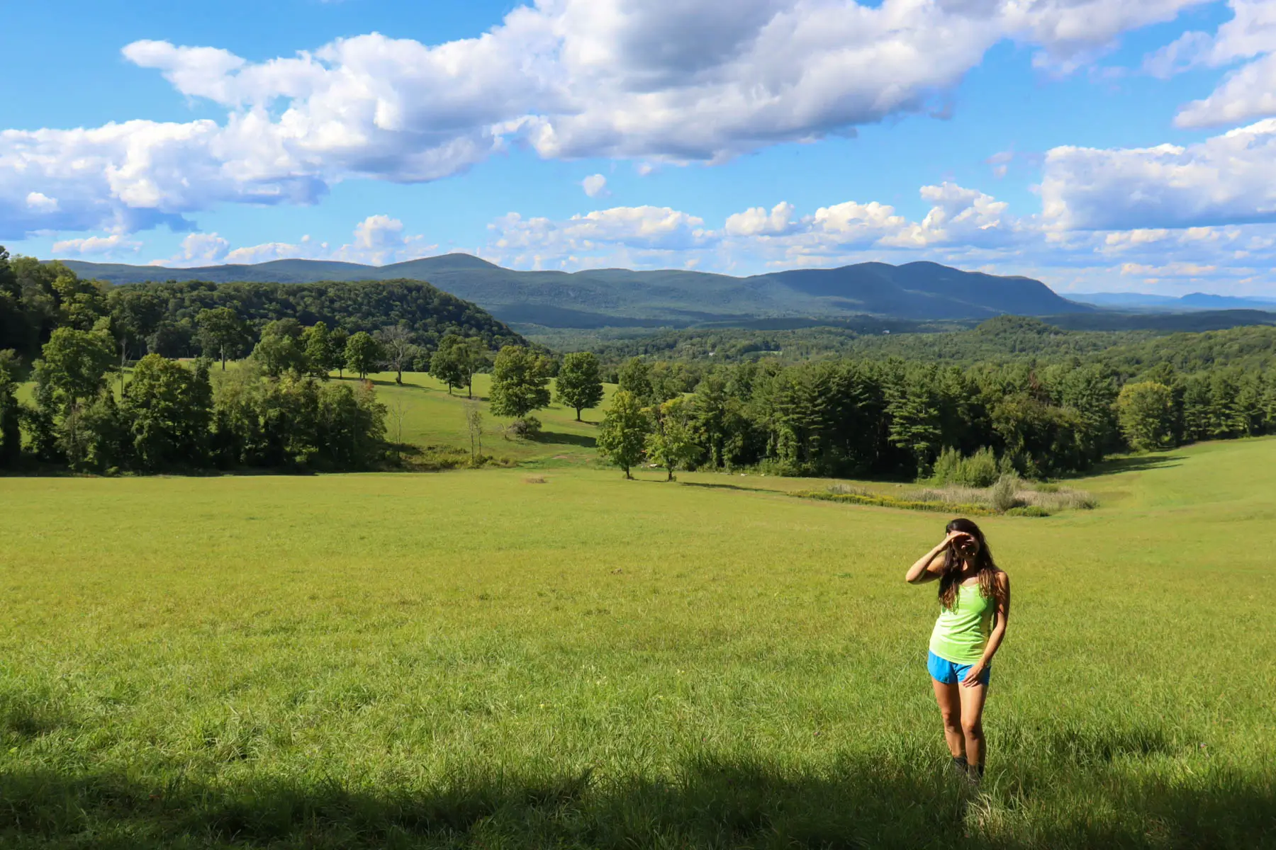 woman in lime green tank top on a grassy open meadow with a view of rolling green hills in the distance and a blue sky with white clouds above.