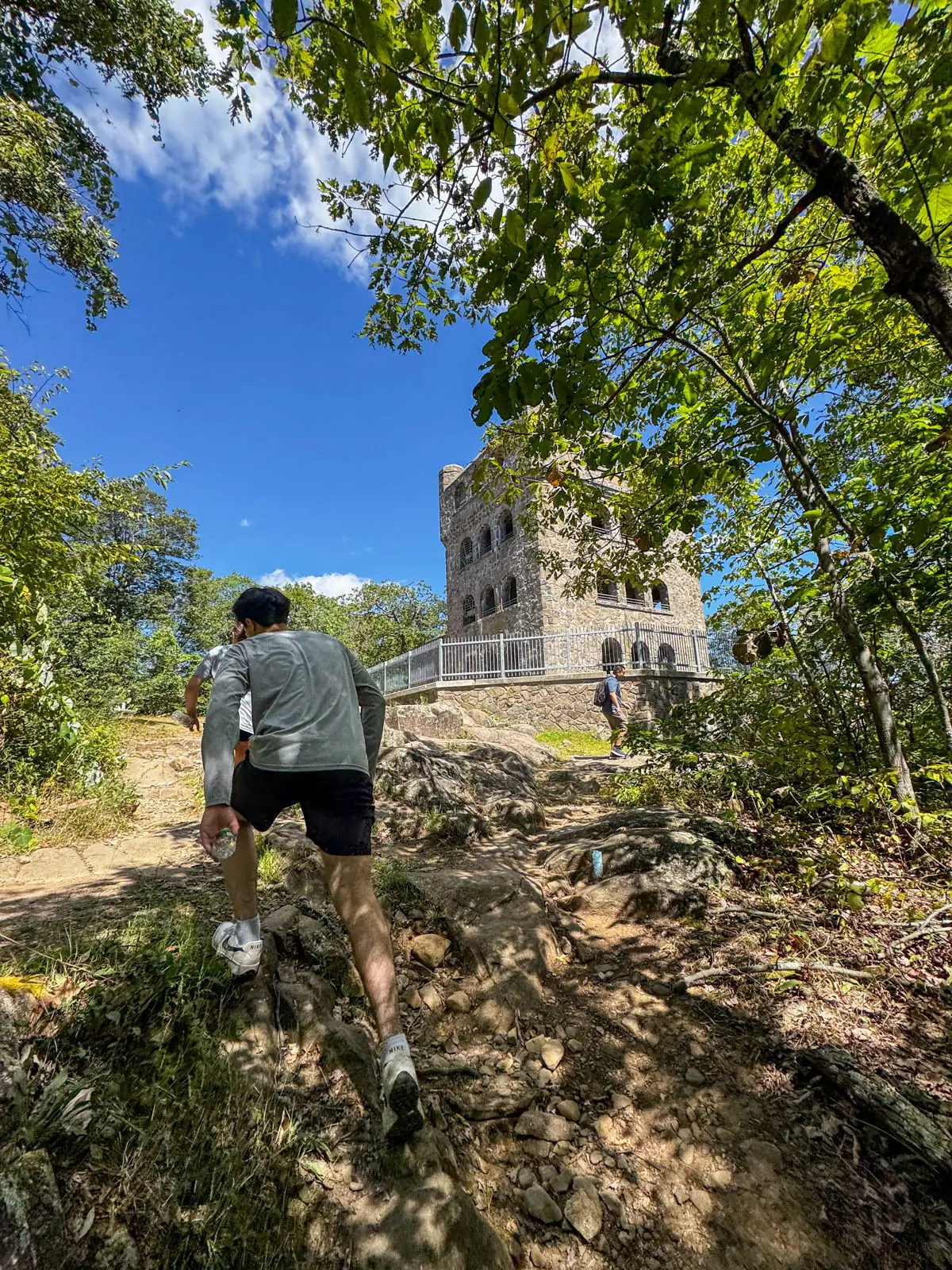 teenage boy walking up rocky path with large stone tower on right with green trees lining the path.