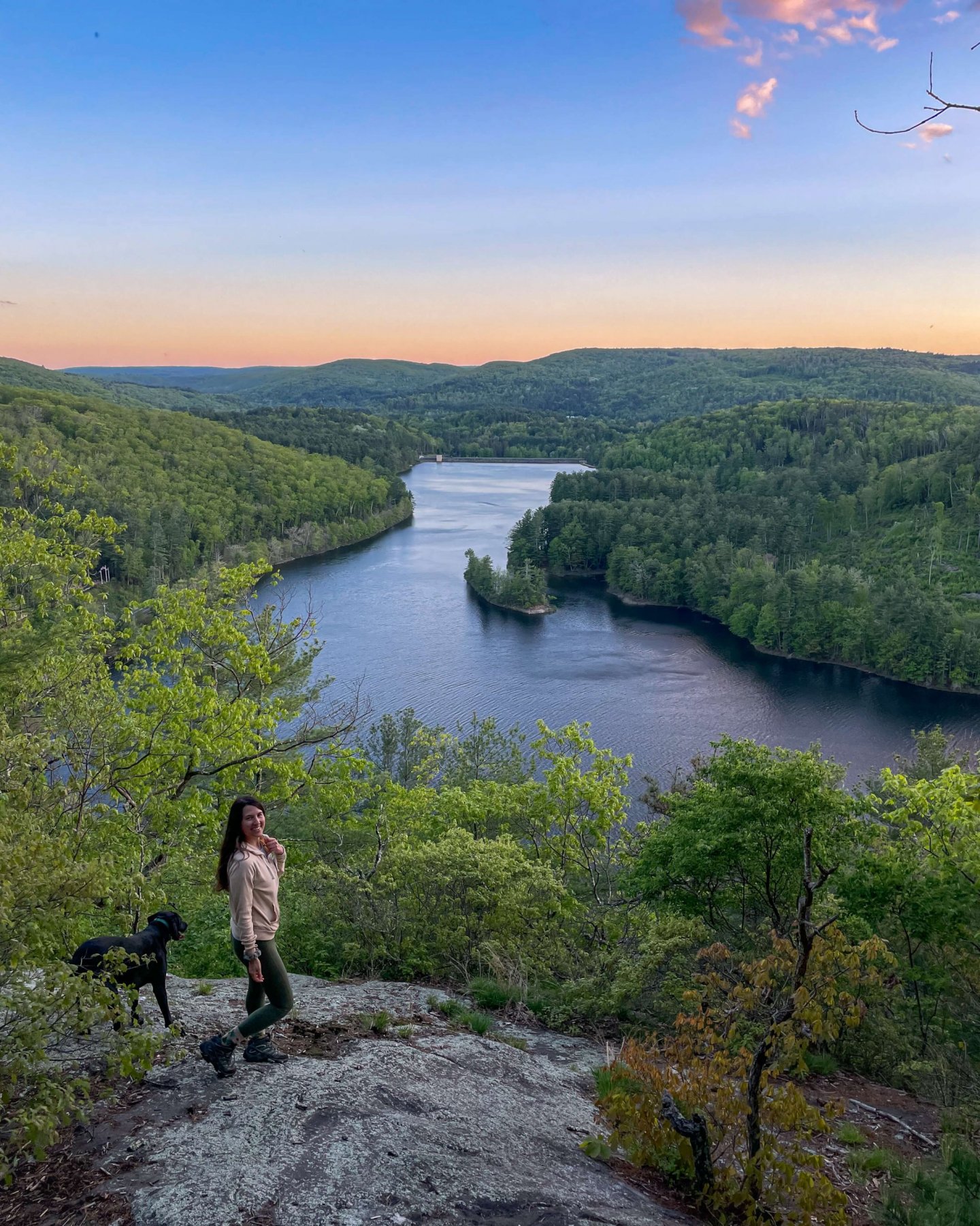 Girl and dog on overlook with the water below, green mountains and sunset in sky.
