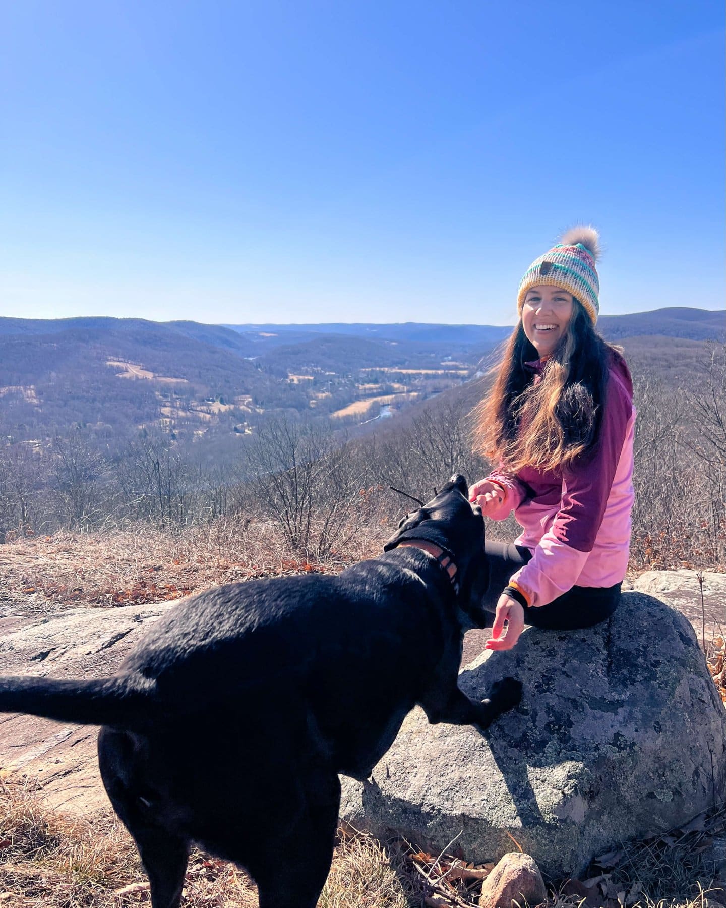 woman in pink shirt and snowcat with black dog on mountaintop