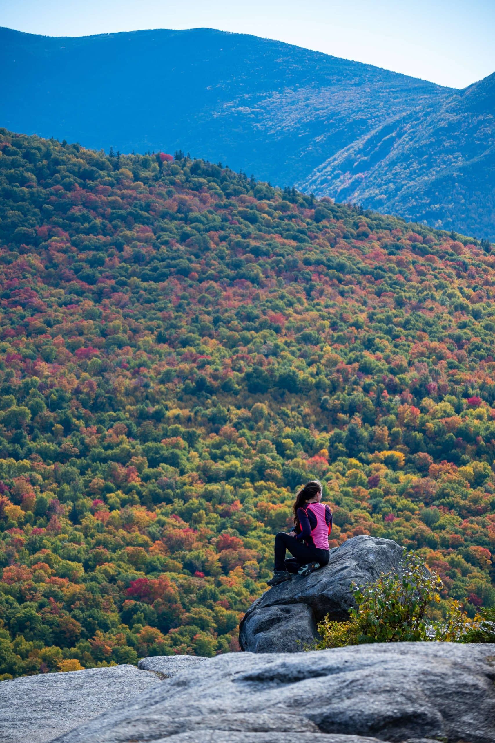 woman sitting on rock on mountaintop in new hampshire with fall colored trees in background.