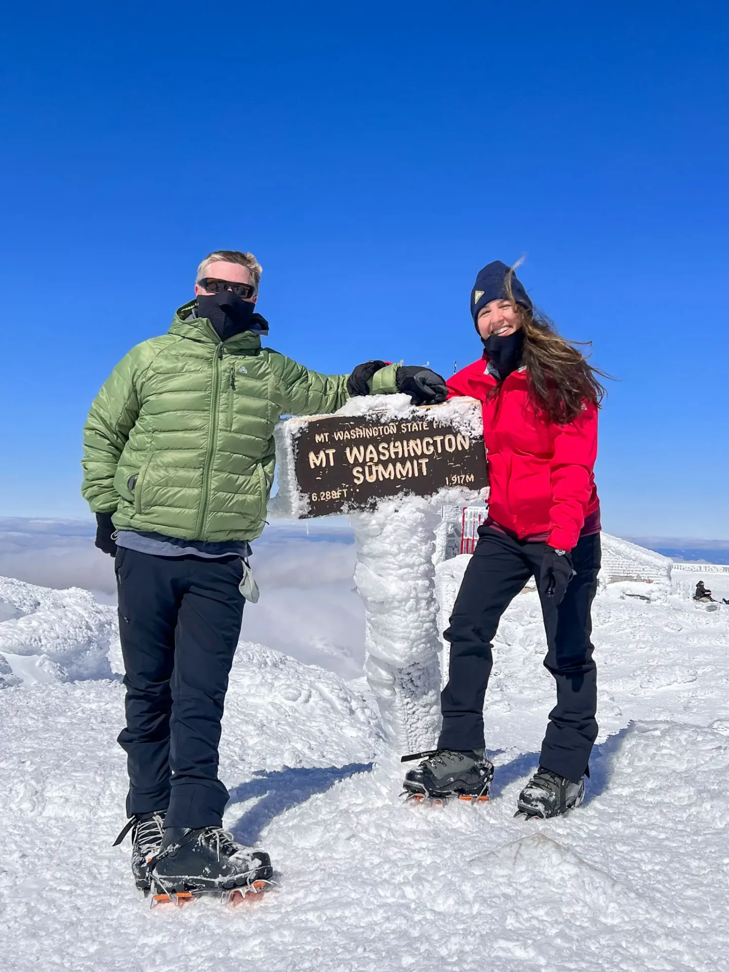 husband and wife in snow gear smiling at top of people walking up a snowy trail to climb mount washington in front of people walking up a snowy trail to climb mount washington sign covered in snow