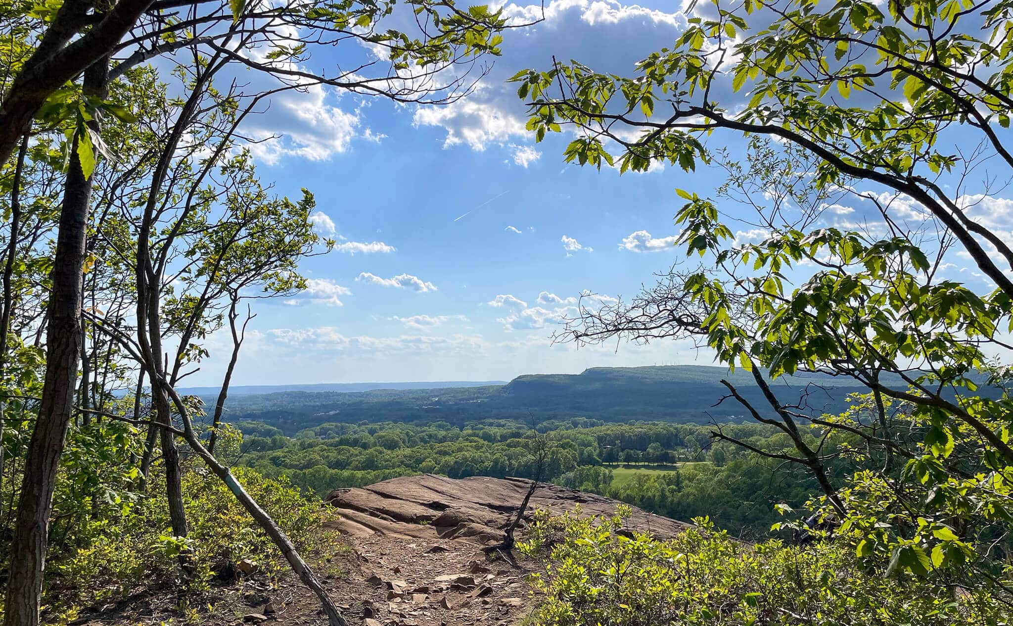 view from cliff in connecticut with green rolling hills in distance and framed with green leaves and trees in summer and blue sky above.