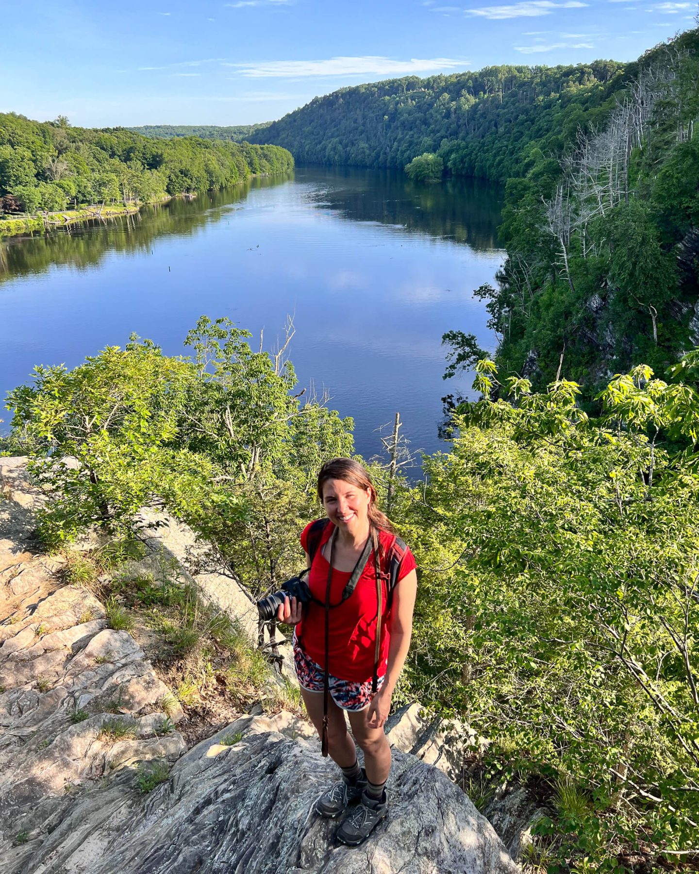 woman in red shirt on summer day on mountaintop with lake behind her and green mountains