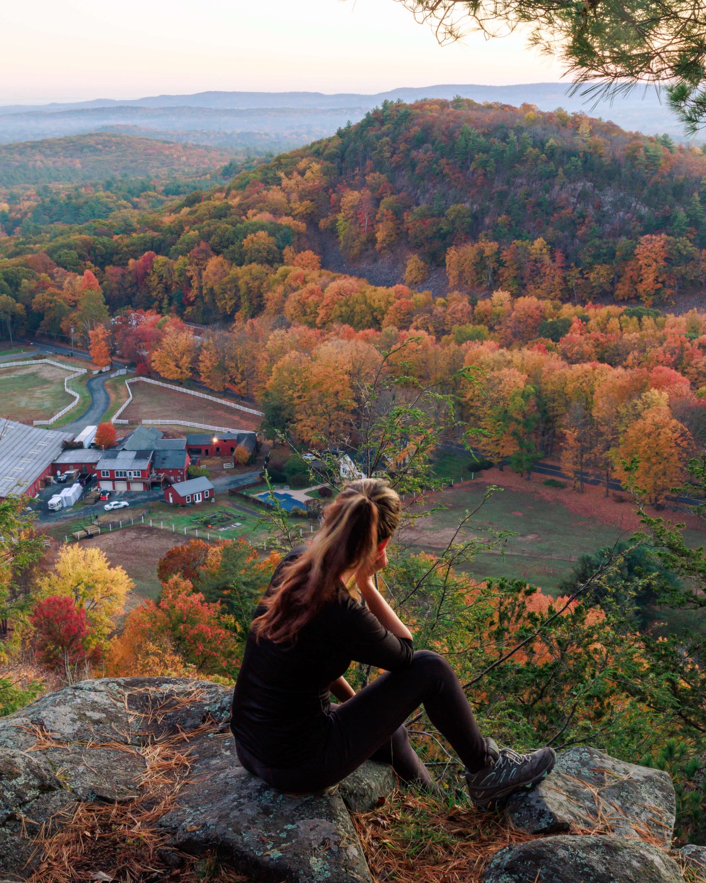 woman dressed in black sitting on mountaintop in fall with orange, red, and yellow trees below.