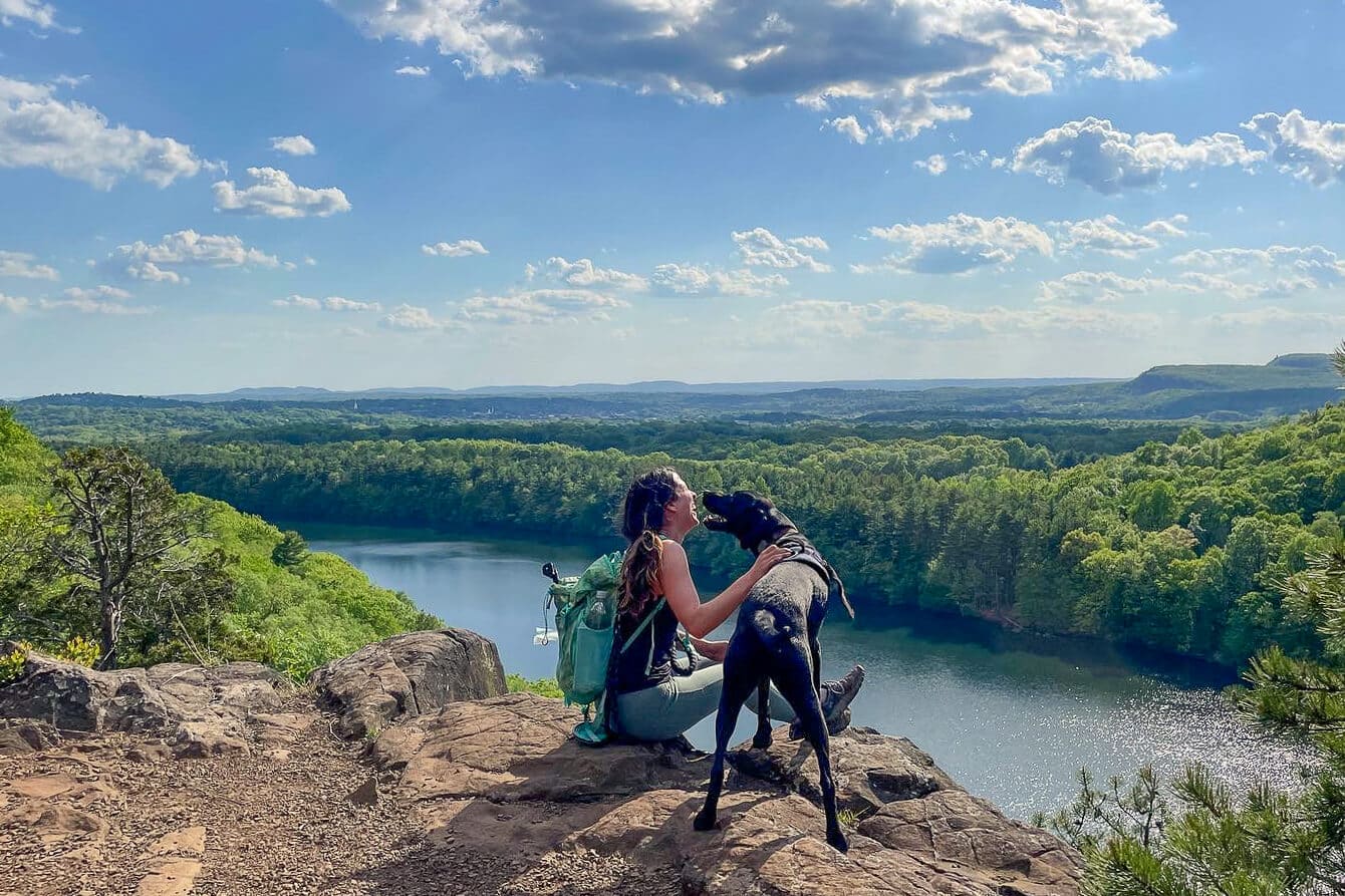 girl and dog laughing on top of mountain hiking with green trees and lake in background