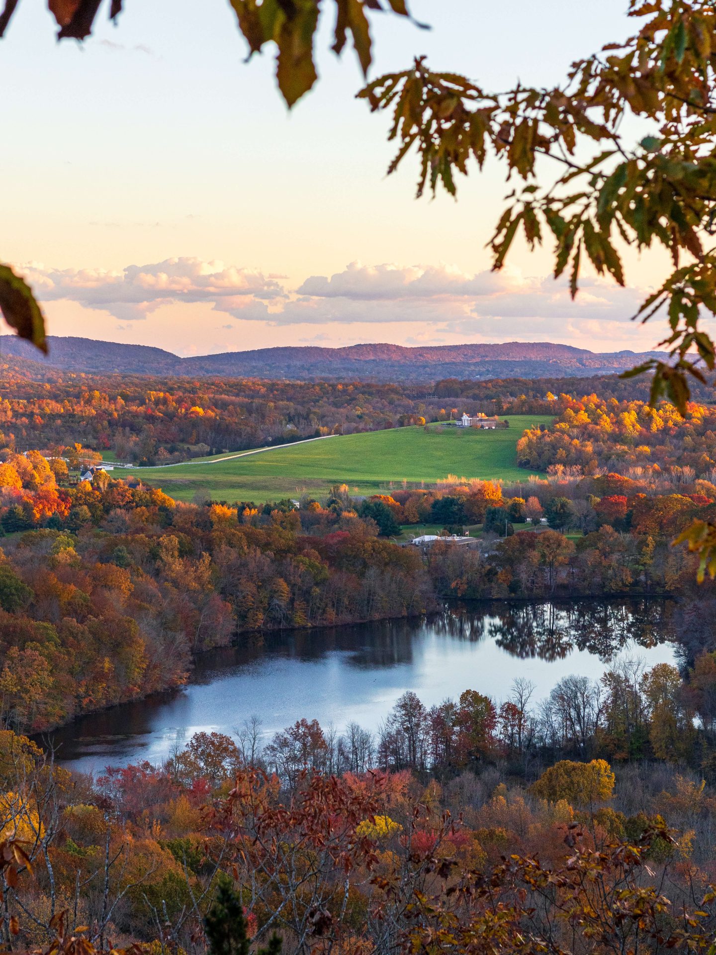 fall view between the leaves on a hike in connecticut with green field and water in distance and orange trees and leaves.