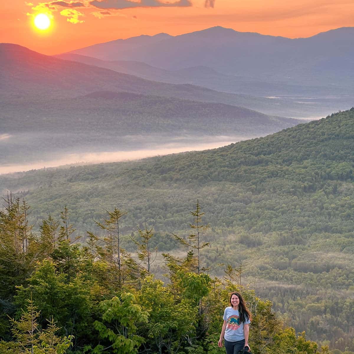 woman on mountaintop in new hampshire with green mountain layers in background and golden sunrise coming up in distance.