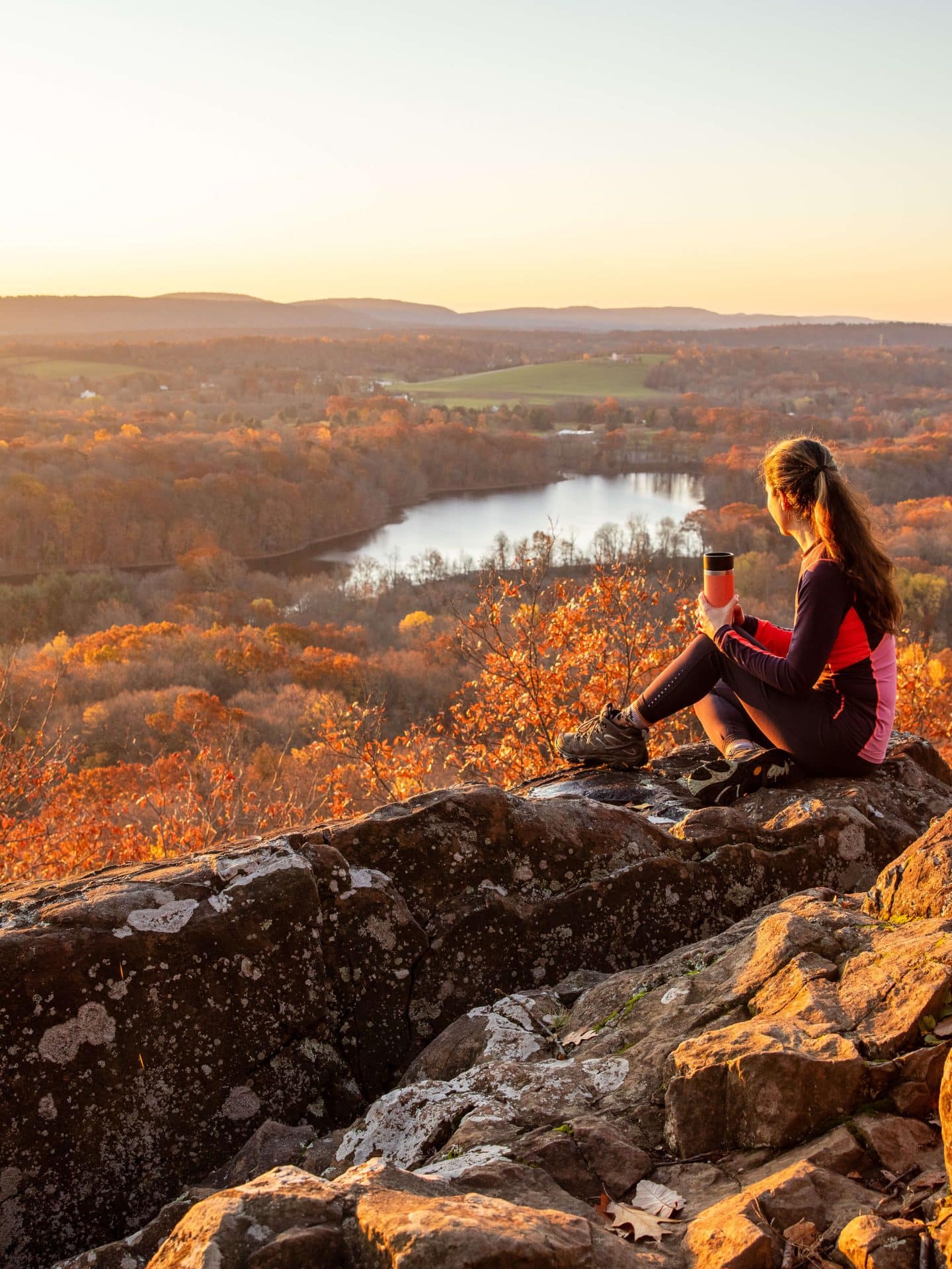 woman sitting on top of cliff looking out at the fall colors holding coffee at sunrise