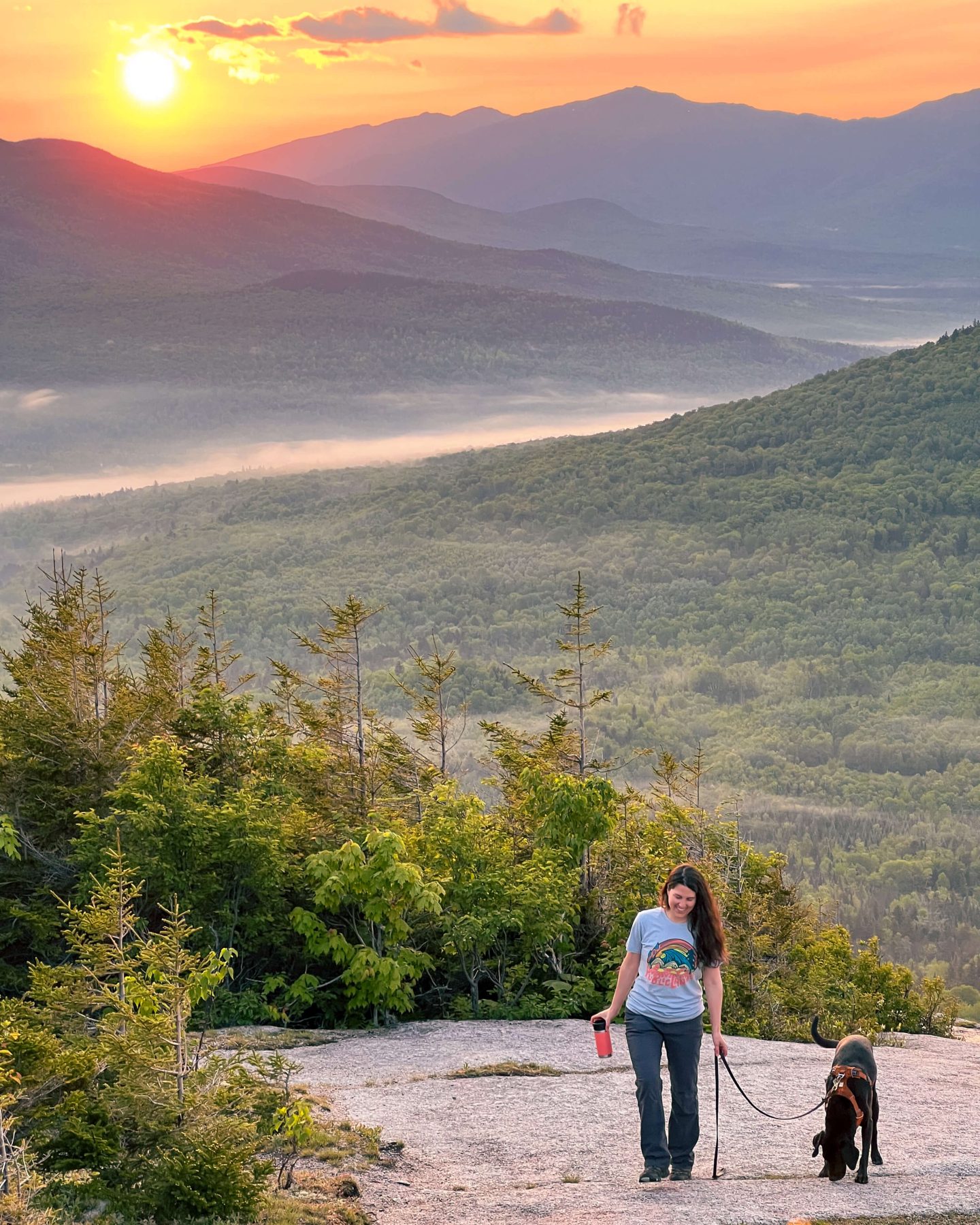 woman and black dog smiling on a sunrise hike in new hampshire, woman holding brown leather leash