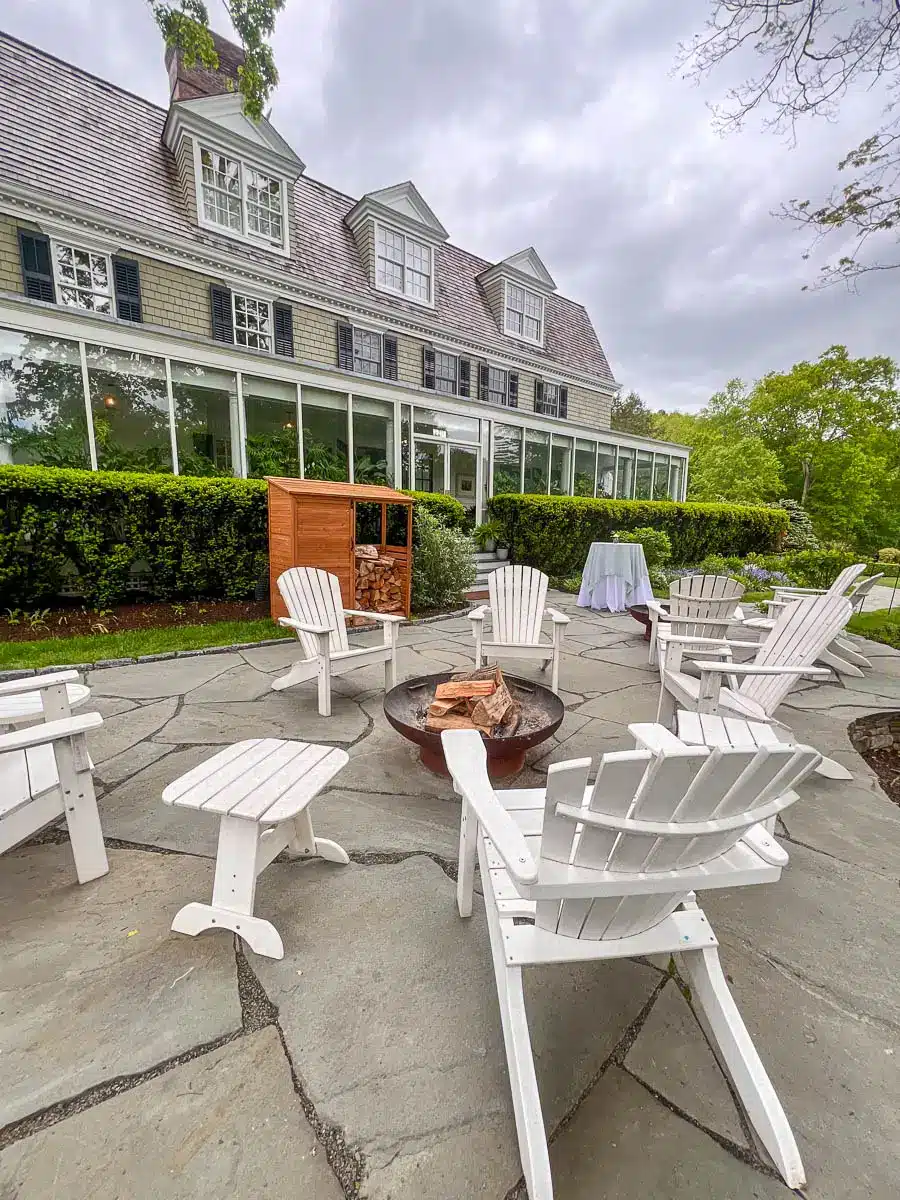 white Adirondack chairs surrounding a fire pit in the daytime outside on a patio in front of big colonial inn.