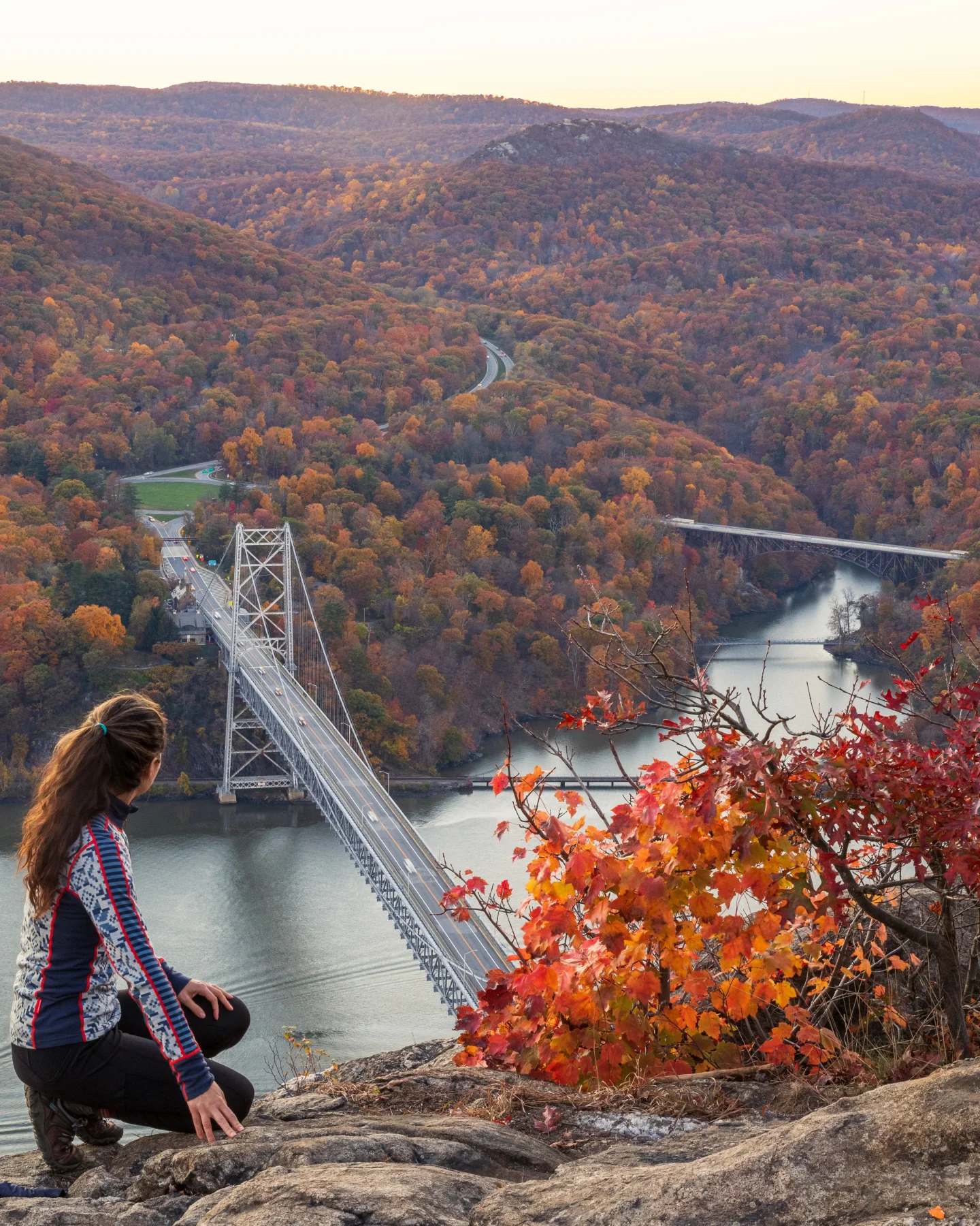 woman sitting on top of anthonys nose hike looking out at bear mountain bridge in fall with orange leave all around