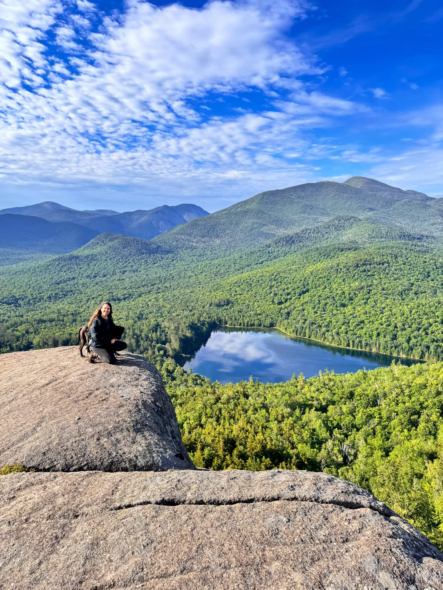 woman smiling with dog on top of mountain in summer in lake placid with green mountains and blue sky