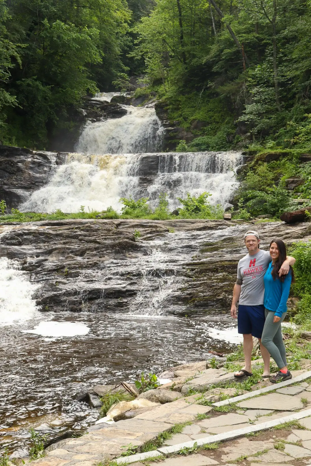 woman in blue shirt and man in gray shirt standing in front of kent falls in summer with green trees all around the waterfall