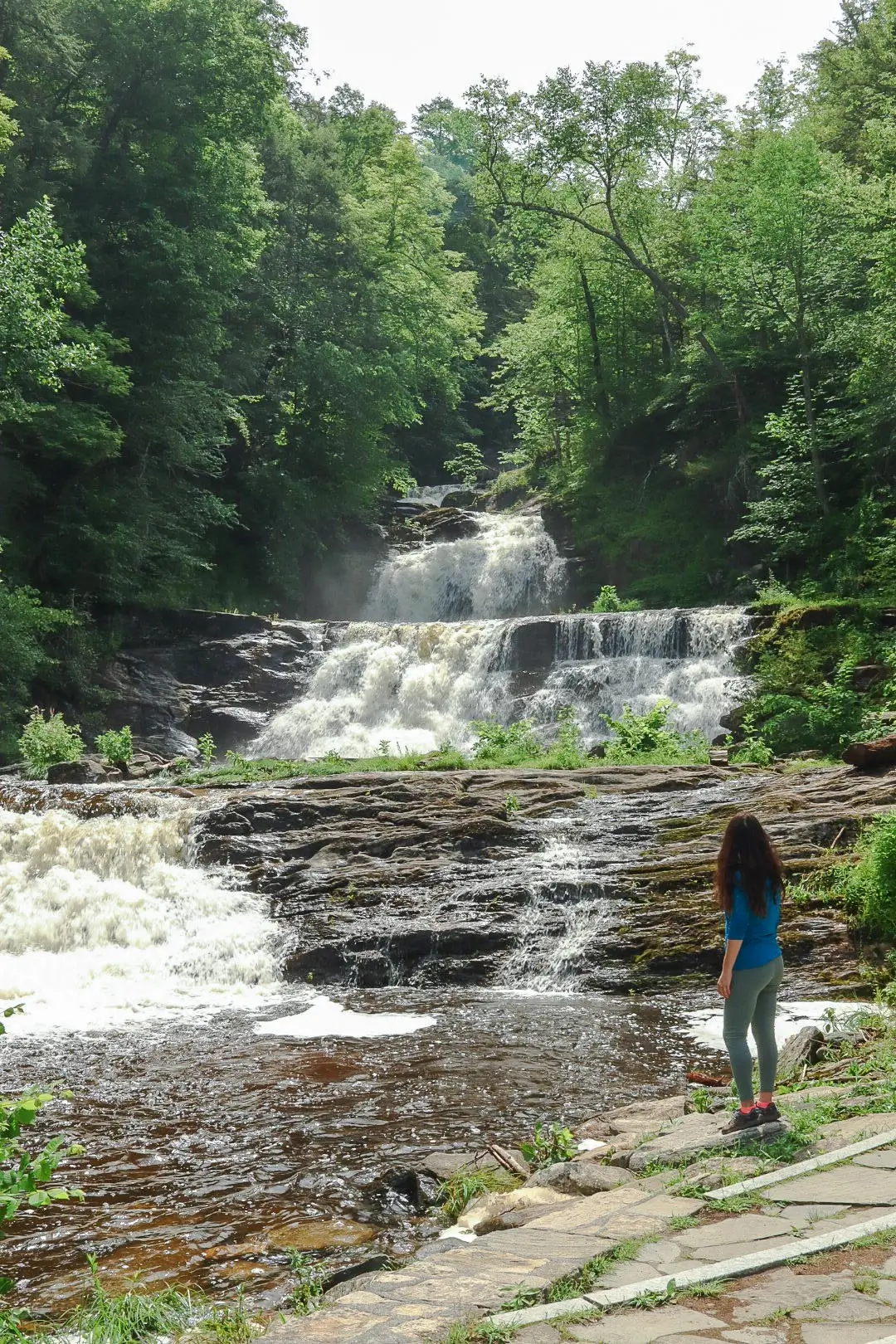 woman in blue shirt standing in front of kent falls in summer with green trees all around the waterfall