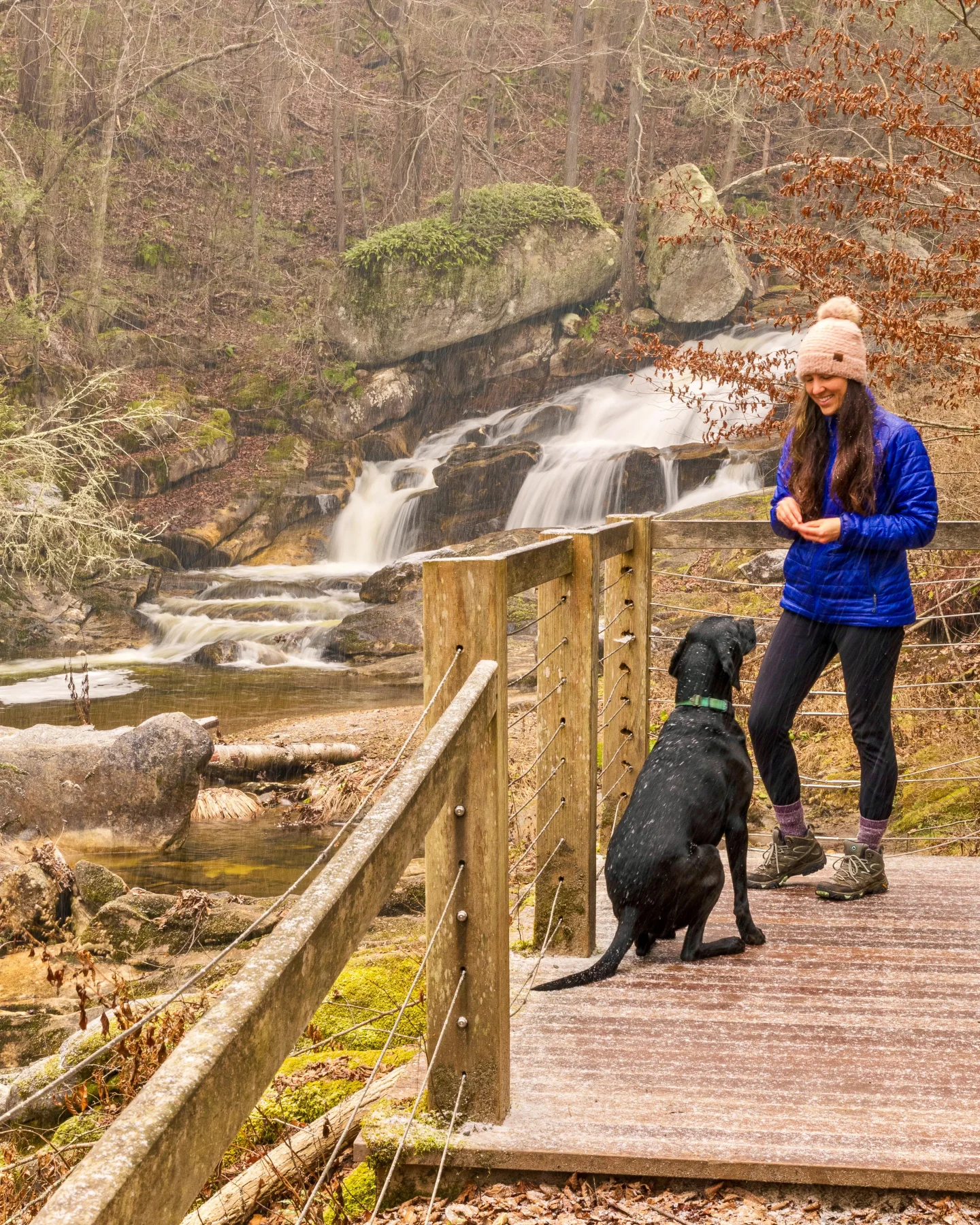 woman in blue winter jacket and pink snow hat and black lab dog in front of kent falls waterfall in winter