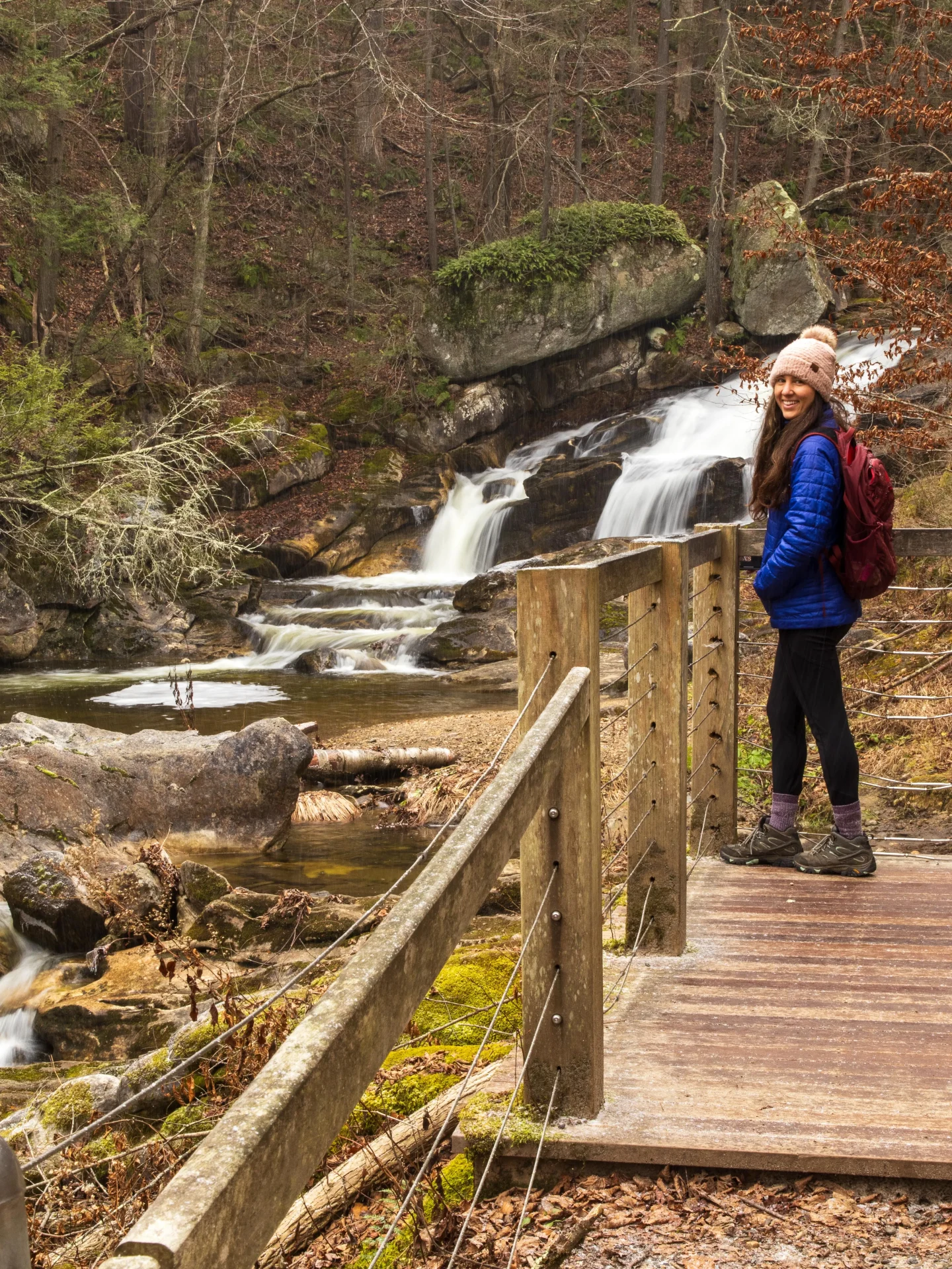 woman in blue winter jacket and pink snow hat and black lab dog in front of kent falls waterfall in winter