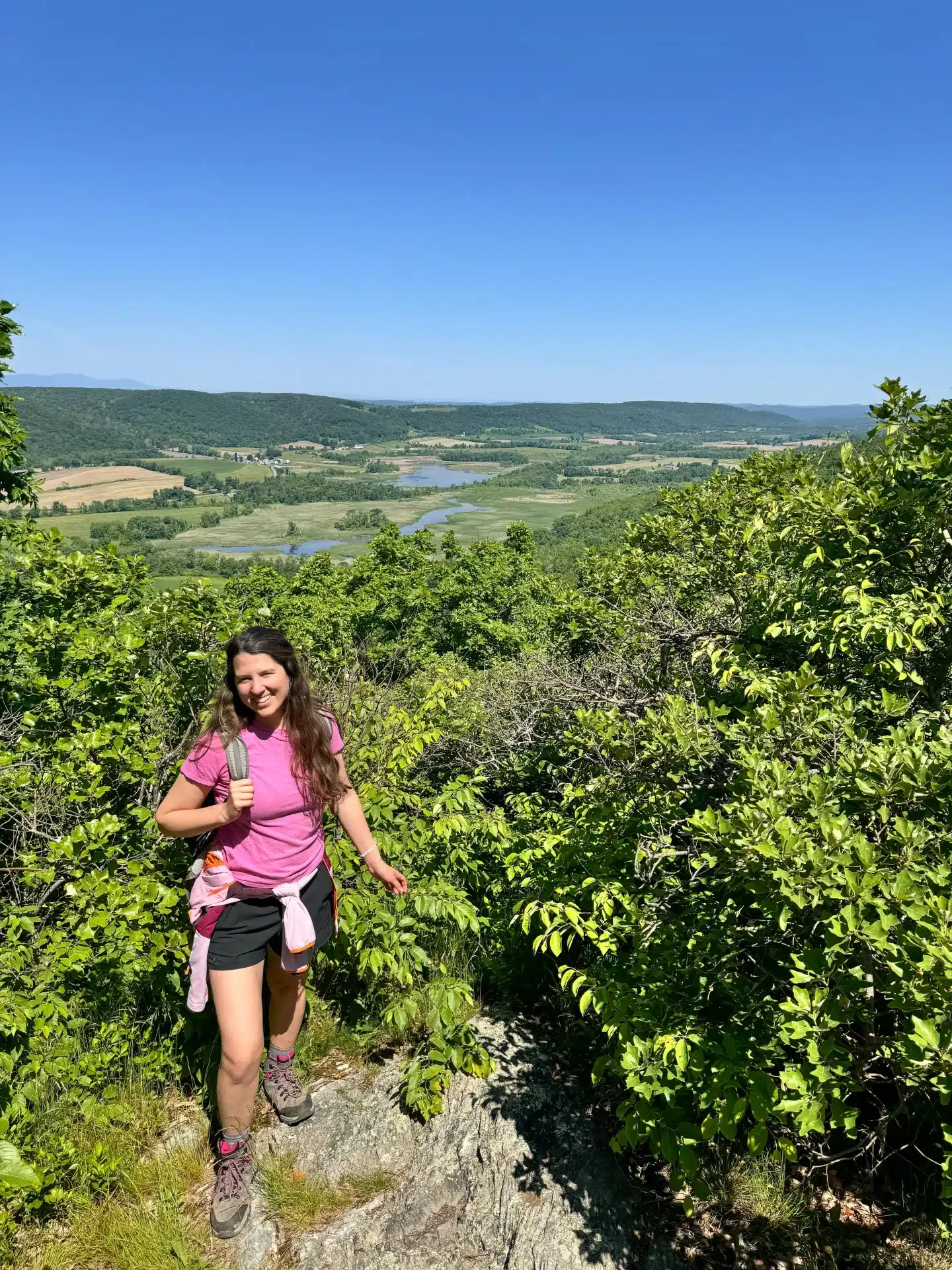 woman smiling in pink shirt looking out at view on top of hike in summer in millerton ny with green trees and mountains in distance