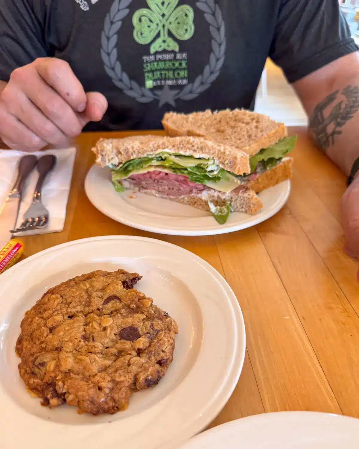 small white round dish with large chocolate chip cookie on it next to another round white dish with a roast beef sandwich on it. 