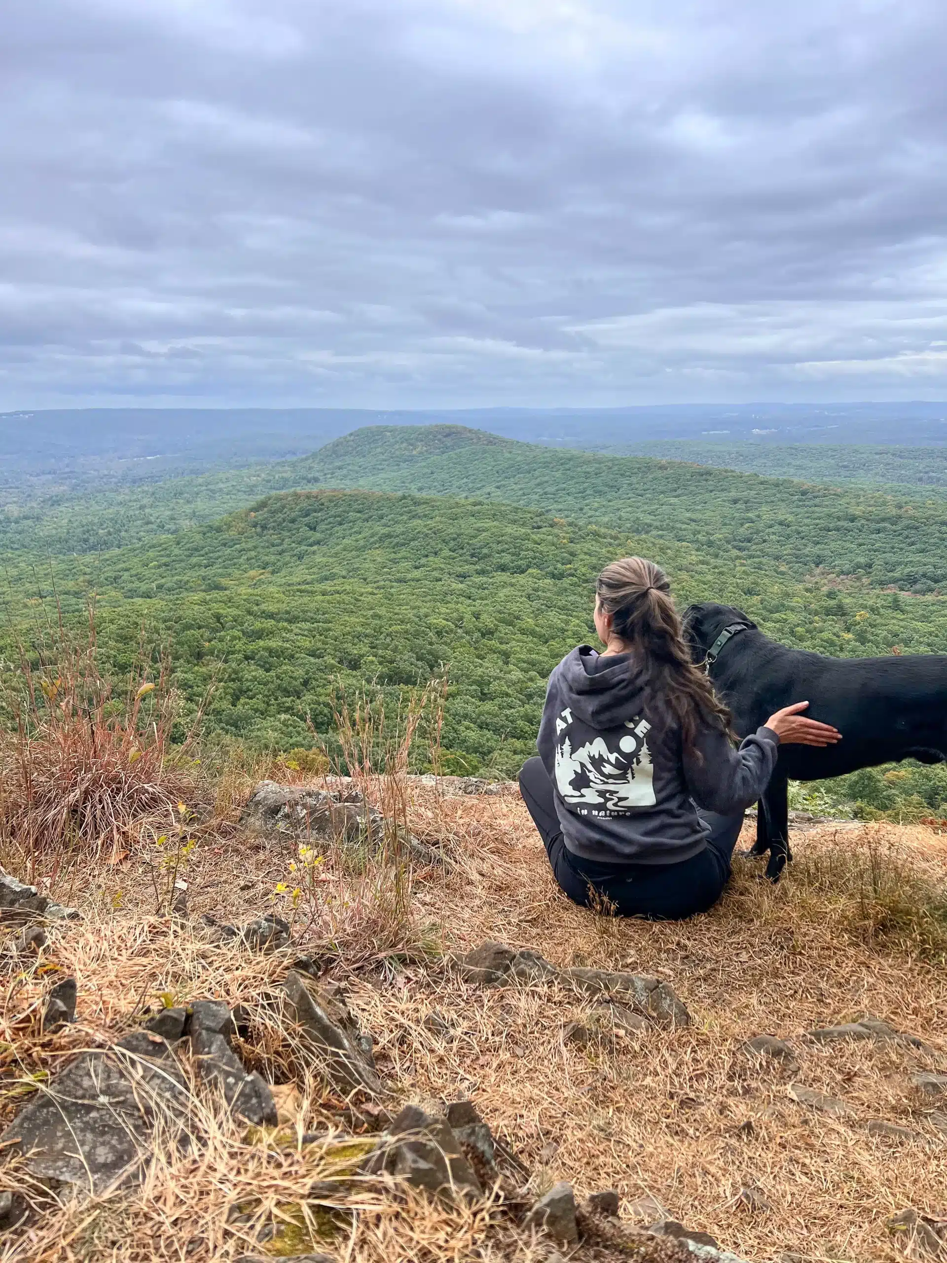 woman smiling on top of mount norwottuck with black dog and green mountains in the background