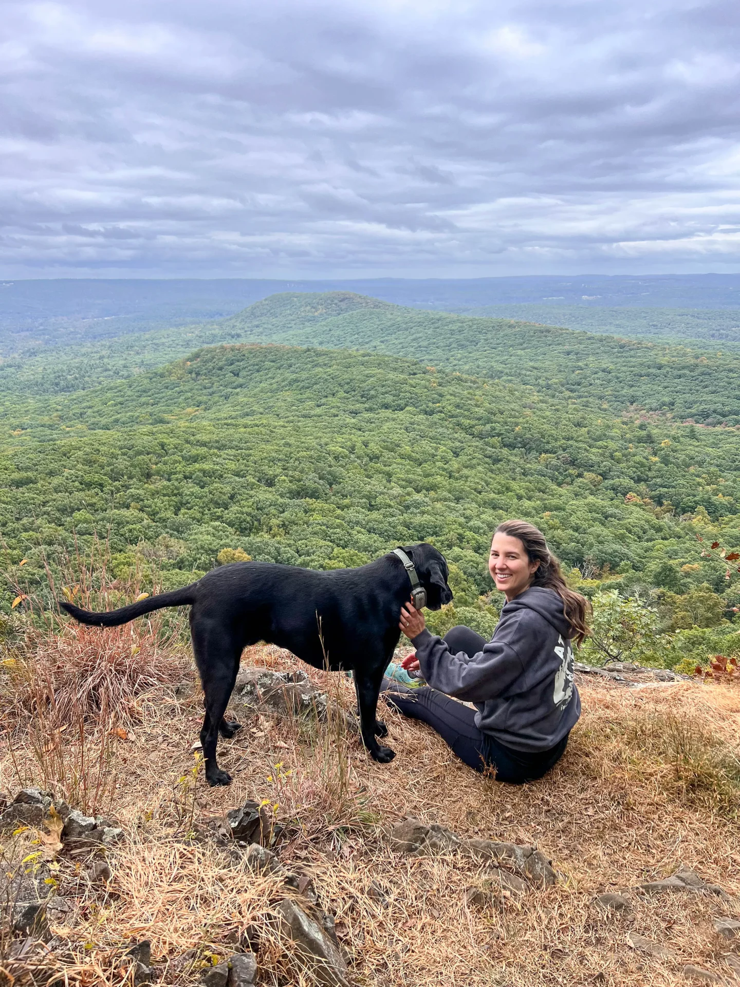 woman smiling on top of mount norwottuck with black dog and green mountains in the background