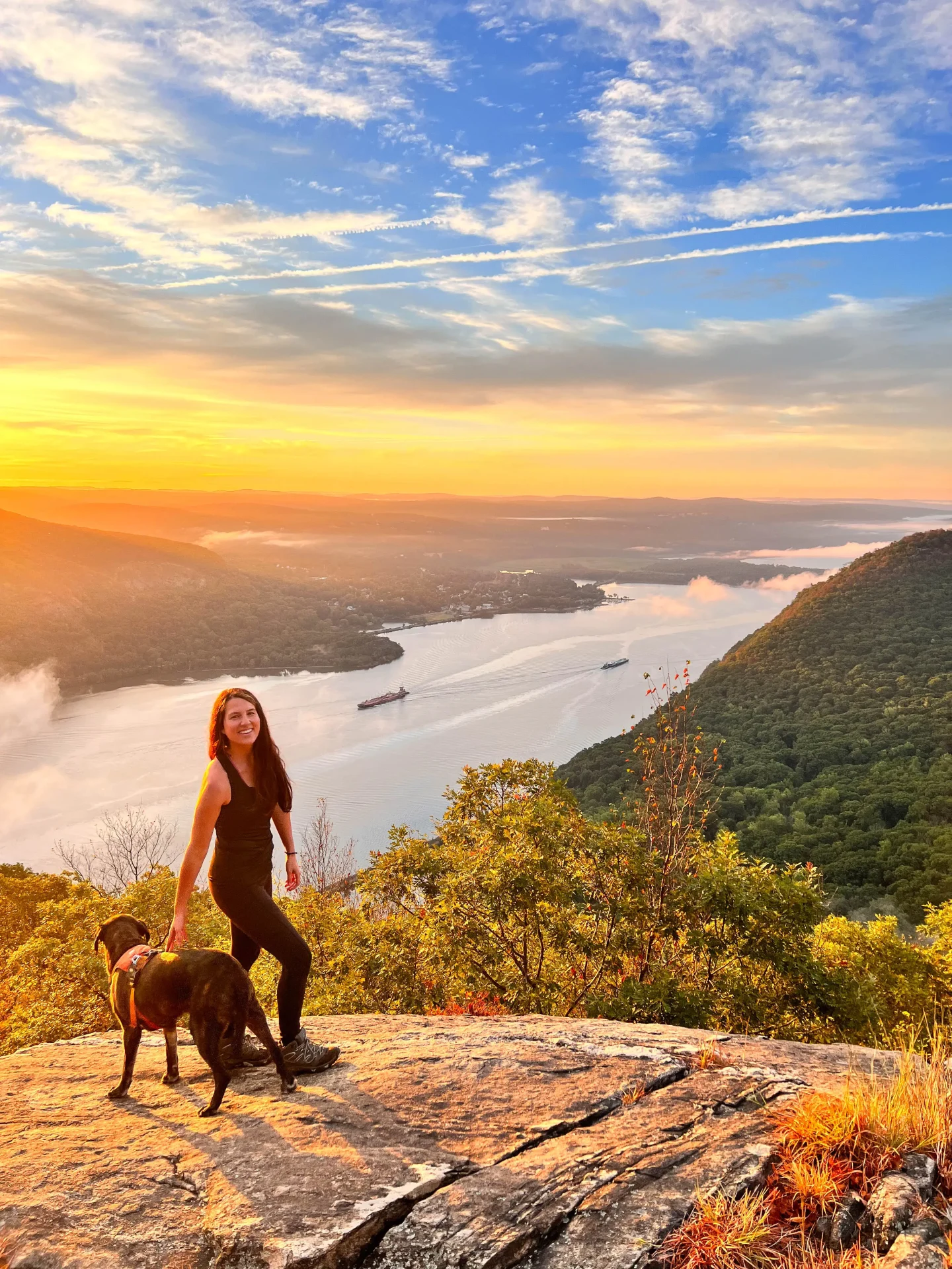 woman in summer in black tank top smiling looking out on early fall sunrise view from top of storm king mountain with orange sun and hudson river below with fog rising