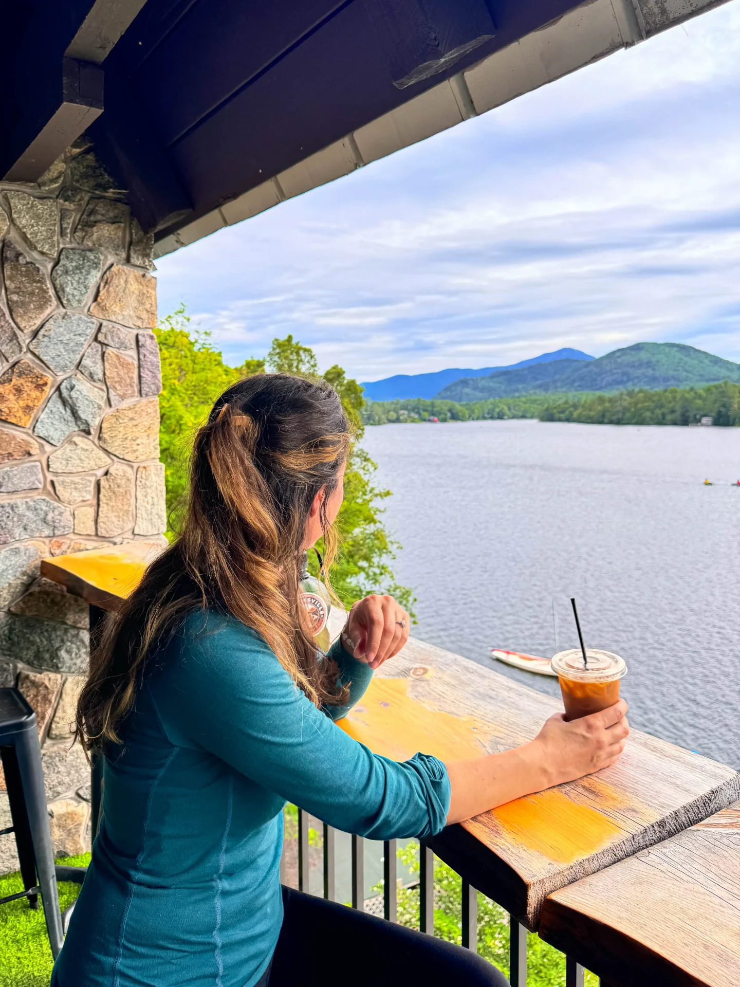 women in blue shirt sitting at the coffee bar in lake placid new york with an iced coffee in hand and view of the lake and mountains