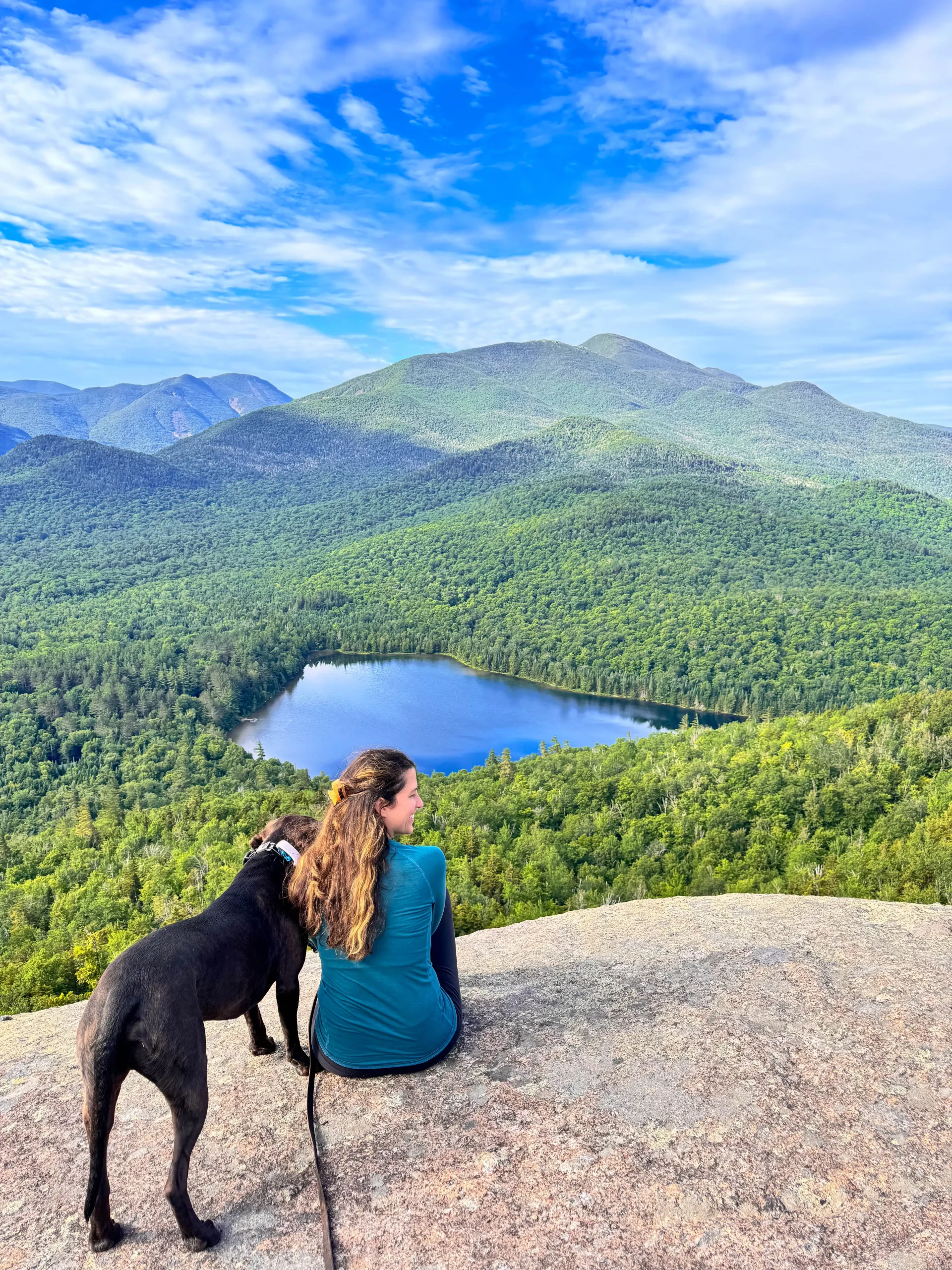 best hike in lake placid woman in blue shirt with brown dog sitting on rock overlooking lake with green mountains all around and blue sky