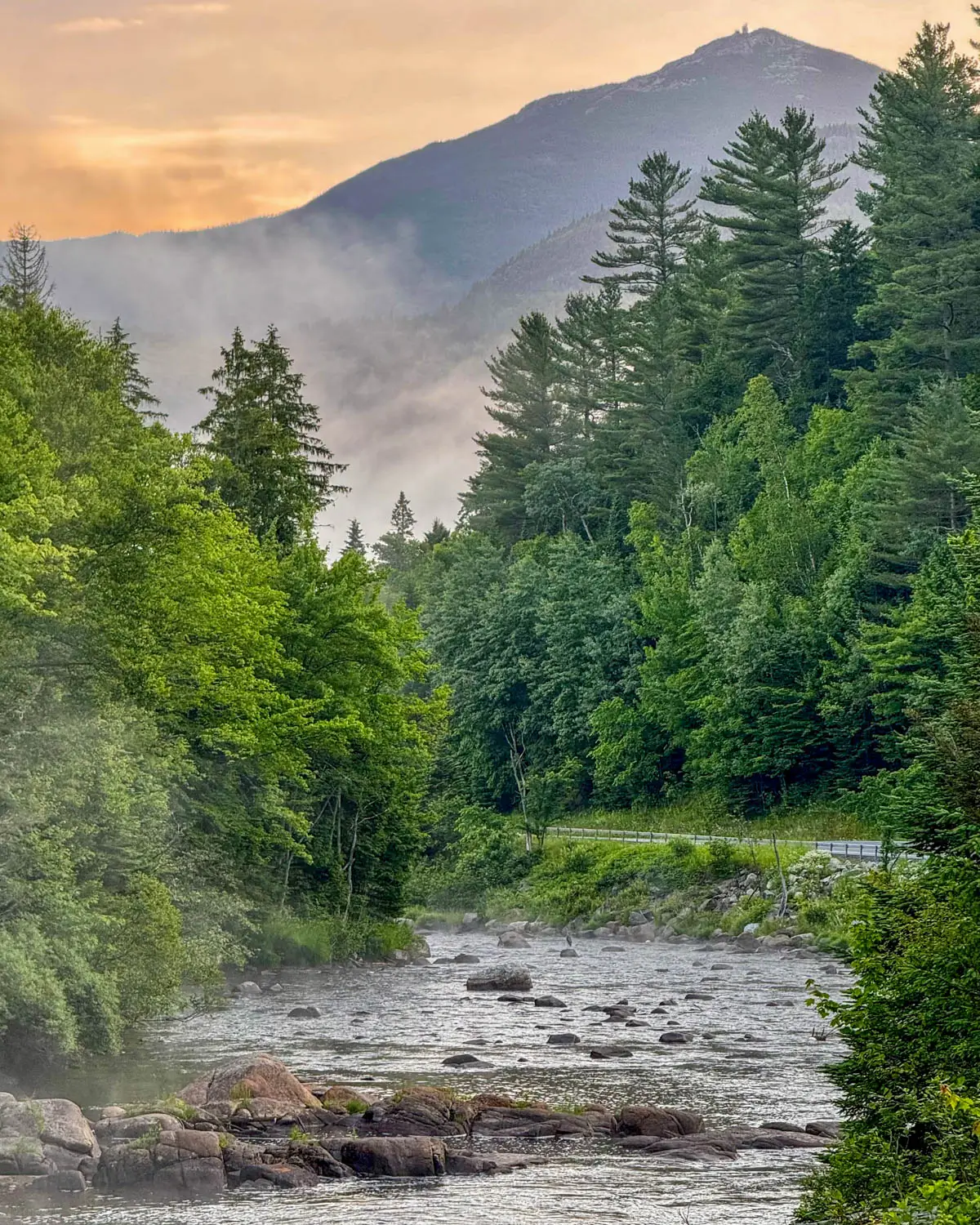 sunrise view of river lined with bright green evergreens and mountain in distance with orange sky outlining it and fog rising up above the river.