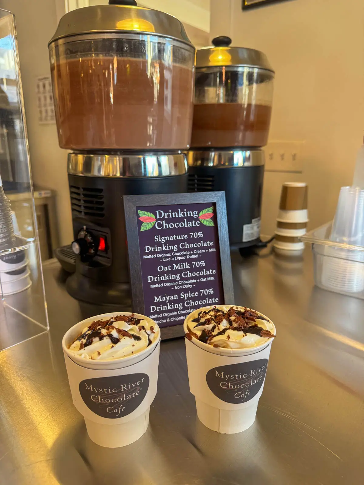 two white cups with whipped cream and chocolate sauce on top at mystic river cafe.