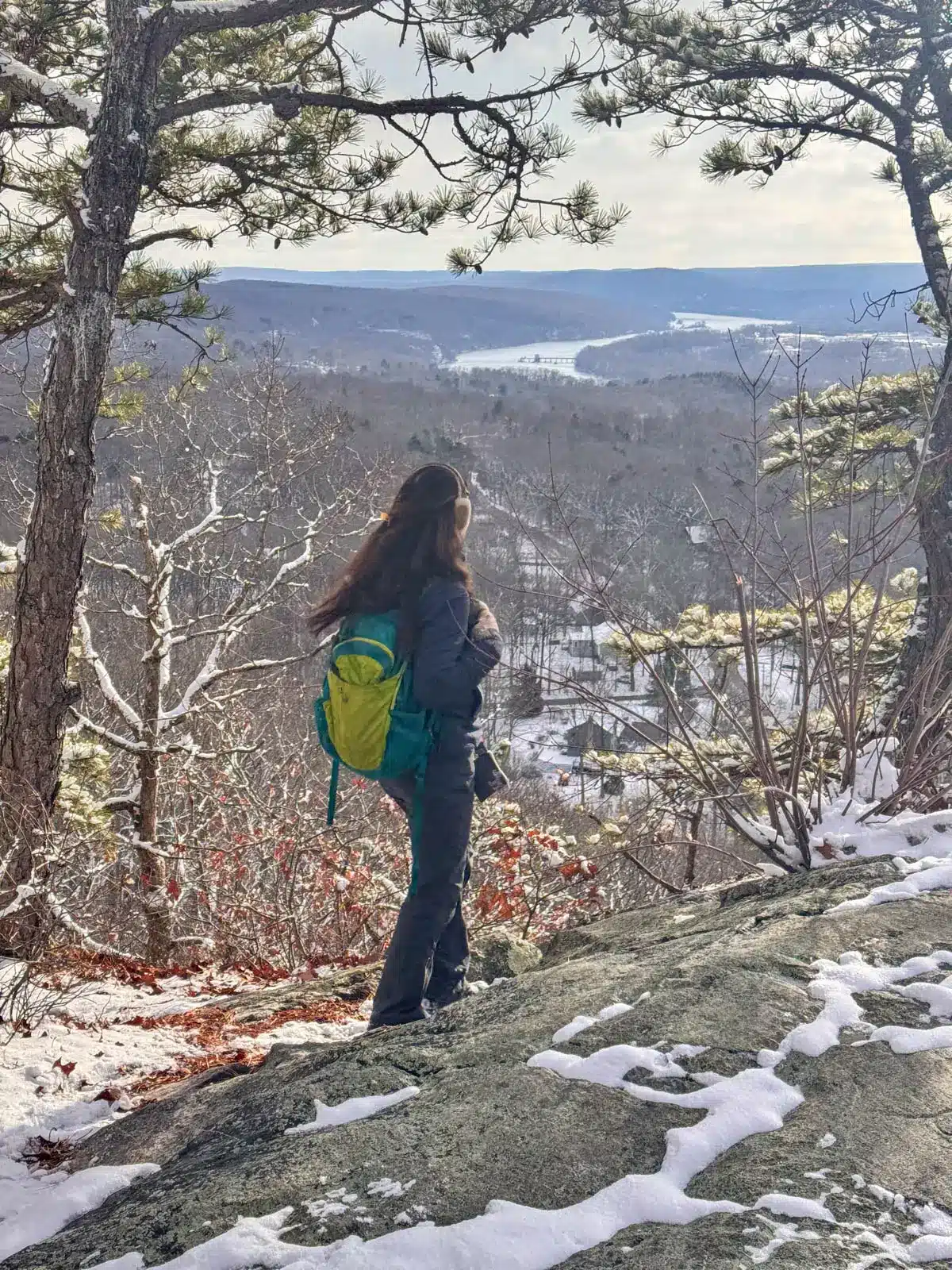 back of brunette woman on winter hike at top of small hill with views of river in the distance.