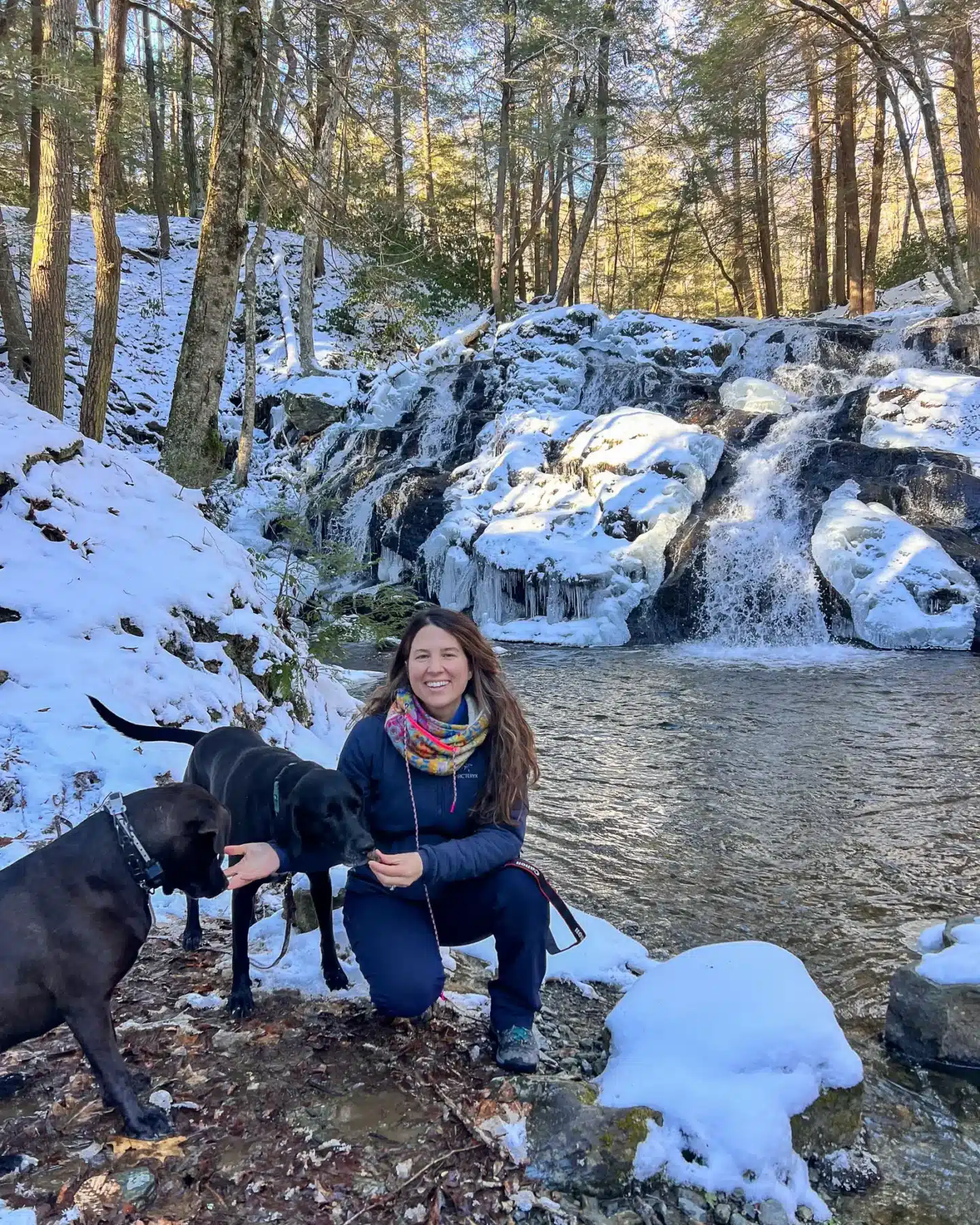 woman crouching down by riga falls with two dogs with waterfall with snow behind them