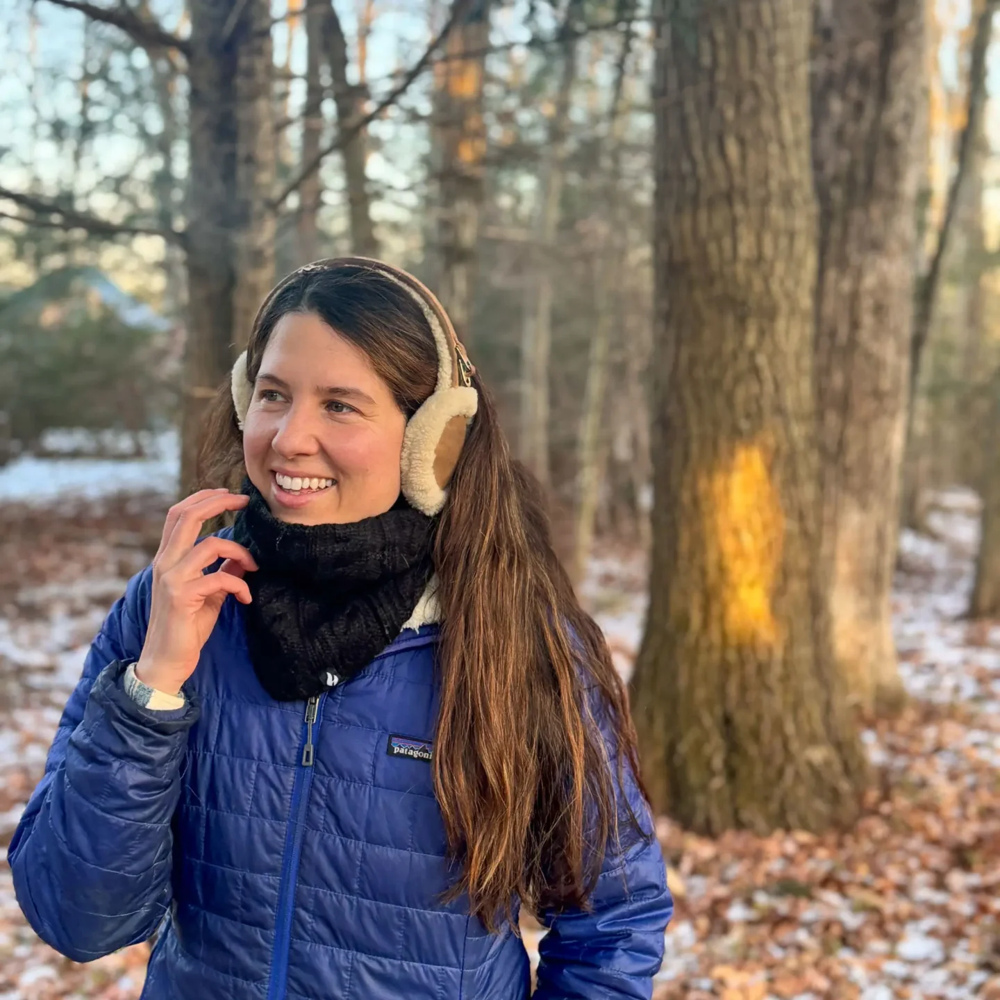 woman in blue winter jacket and beige earmuffs smiling on a winter walk wearing a black neck gaiter