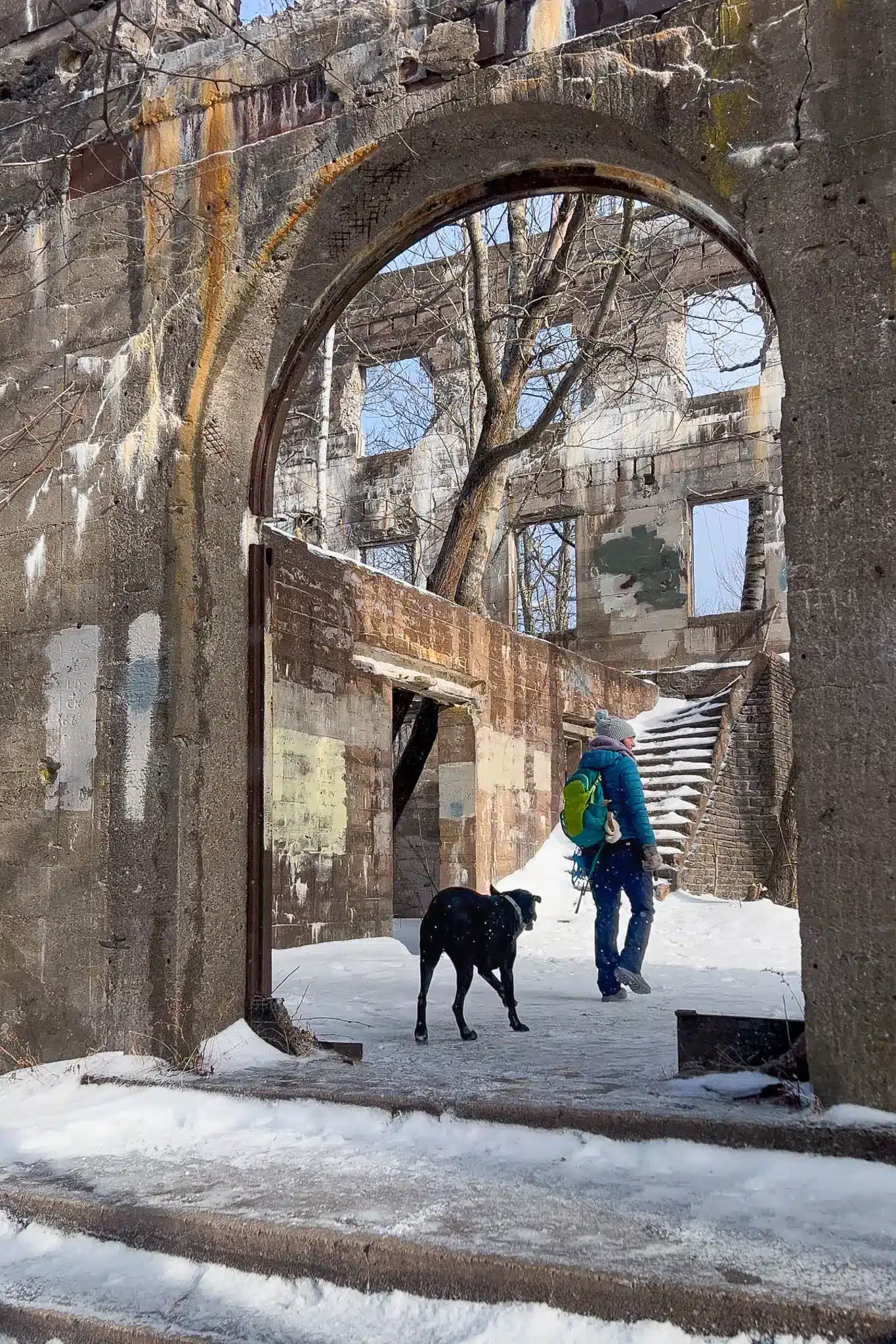 woman with blue winter jacket and bright green backpack walking through stone archway of overlook mountain house in catskills new york with snow on the ground