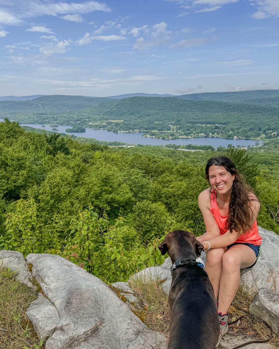 brown dog and woman with brown hair in coral tank top smiling at the camera at the top of cheshire cobbles hike with rolling green mountains in the distance