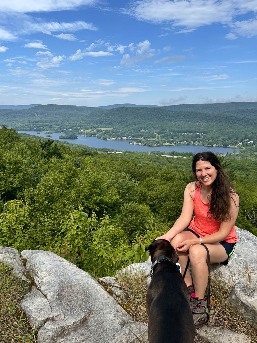 brown dog and woman with brown hair in coral tank top smiling at the camera at the top of cheshire cobbles hike with rolling green mountains in the distance