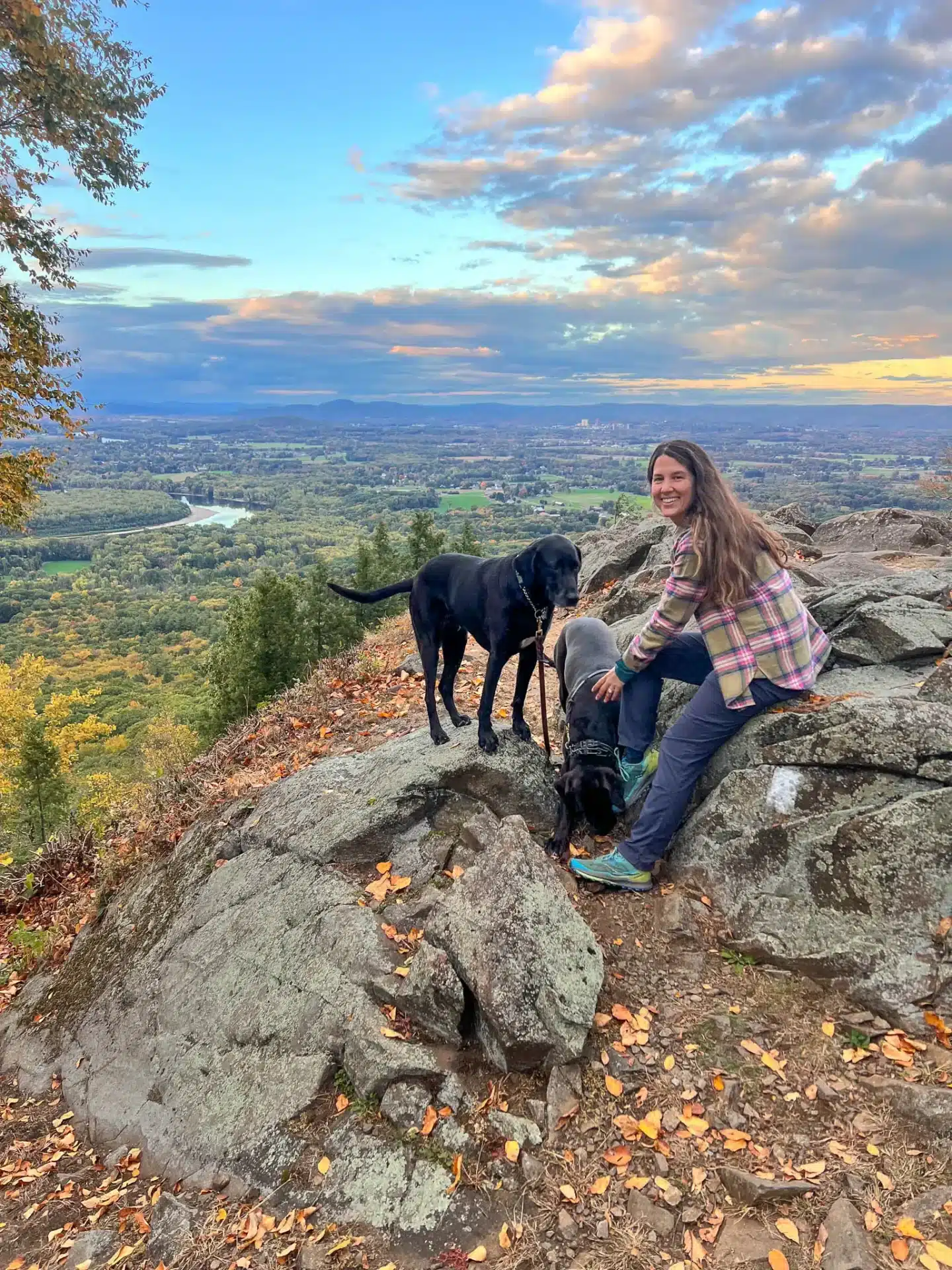 woman in flannel smiling with black dog on summit of mount holyoke in early fall with green mountains and light yellow leaves in the distance