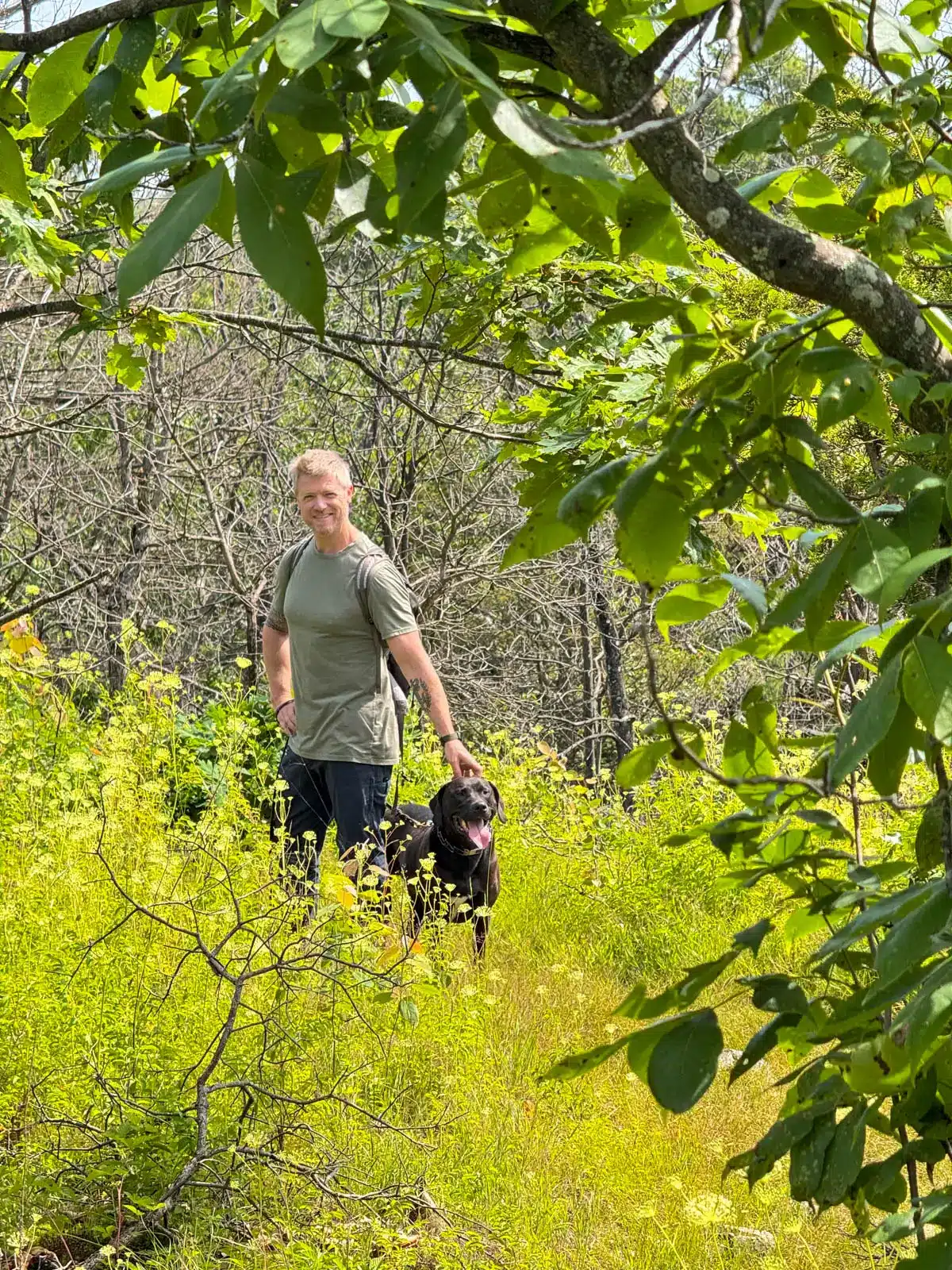 man standing in bright green grass on a hike with brown dog smiling at the camera.