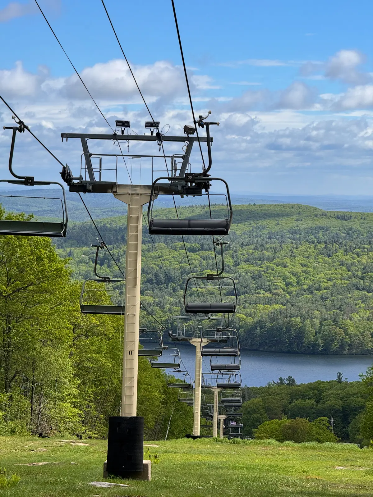 view of ski lift in spring along a loop trail at mount wachusett with bright green mountains and blue sky in the distance.