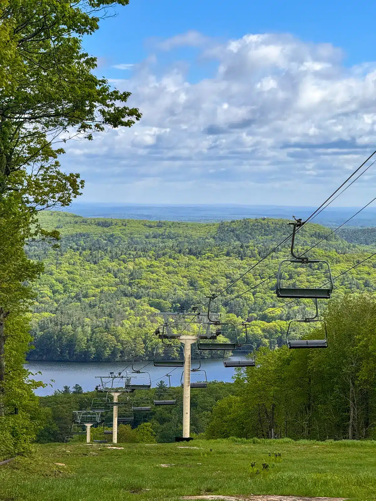 view of ski lift in spring along a loop trail at mount wachusett with bright green mountains and blue sky in the distance.