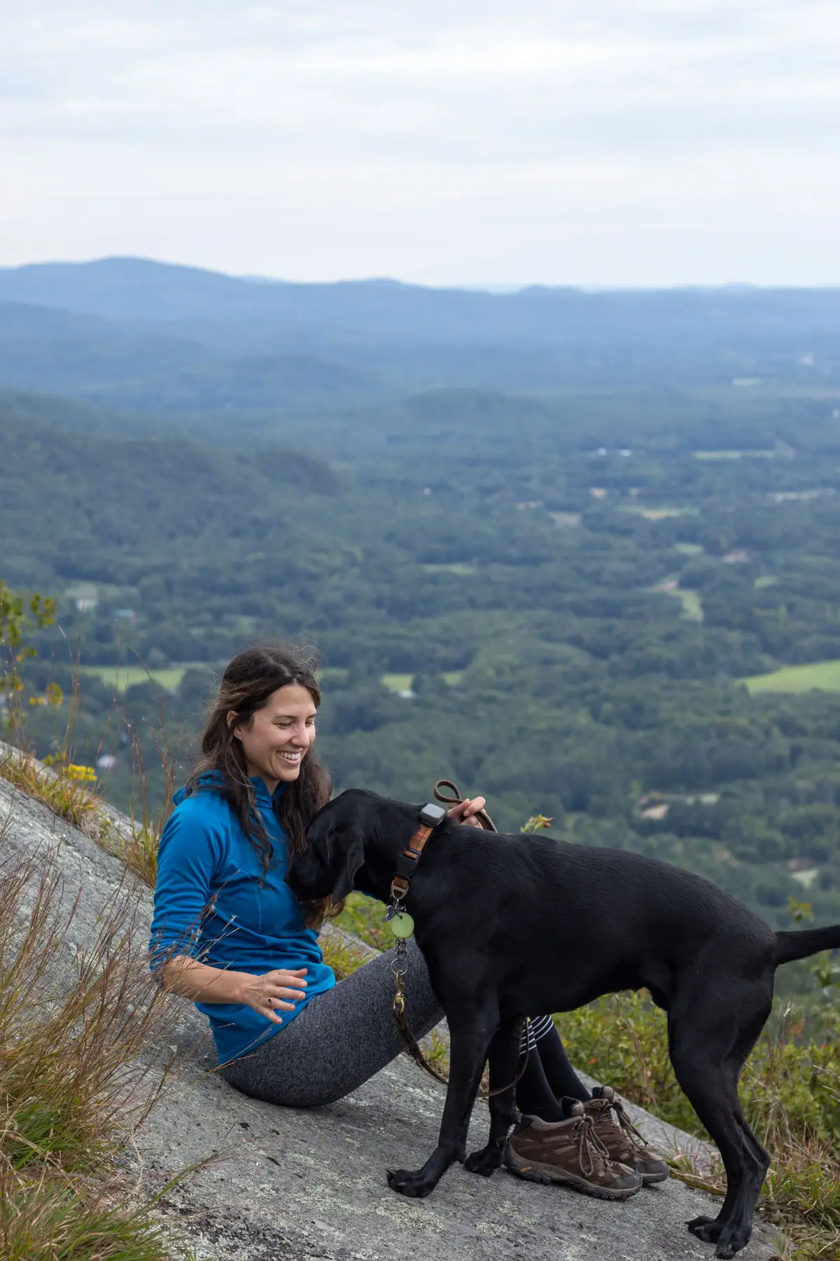 woman smiling on mountain summit in blue shirt petting black dog with green new hampshire mountains in the background.