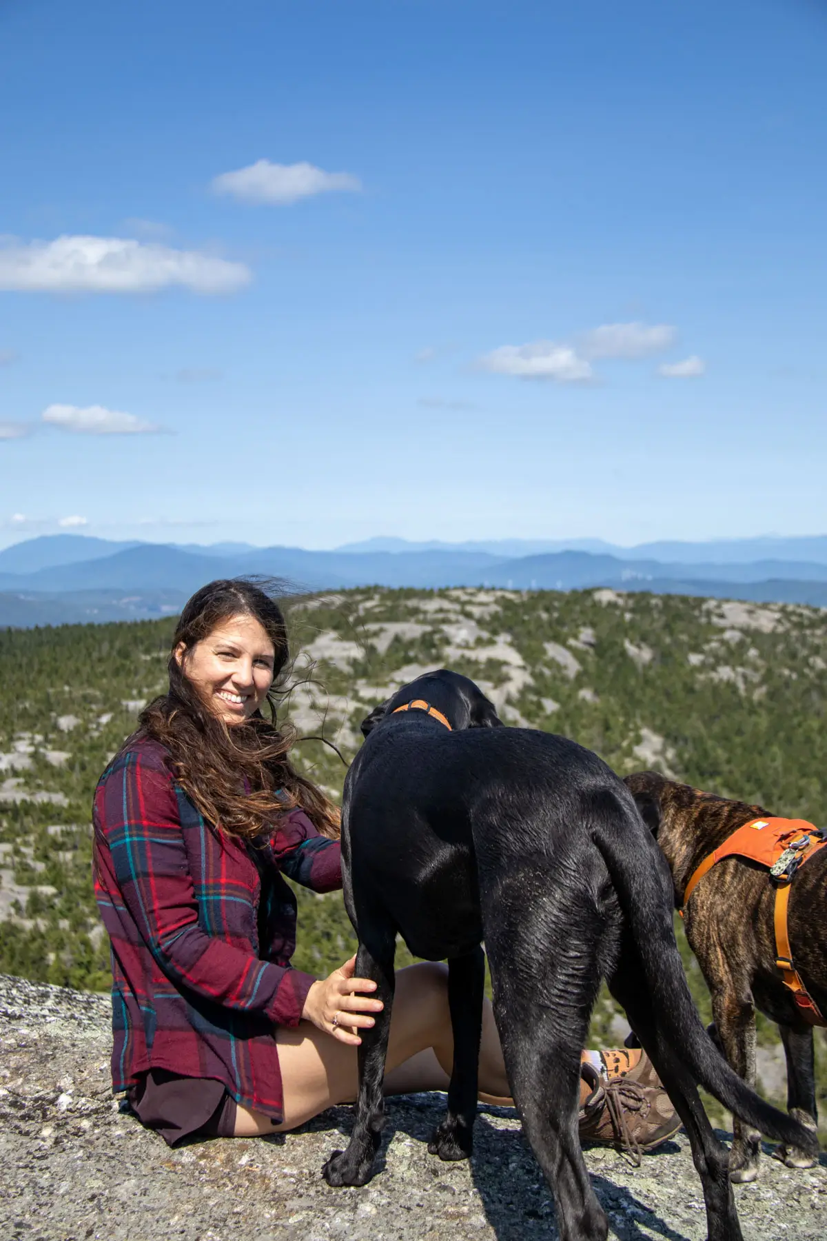 woman in maroon flannel smiling at the camera with black dog on exposed rocky summit in new hampshire with green mountains behind him and blue sky above.