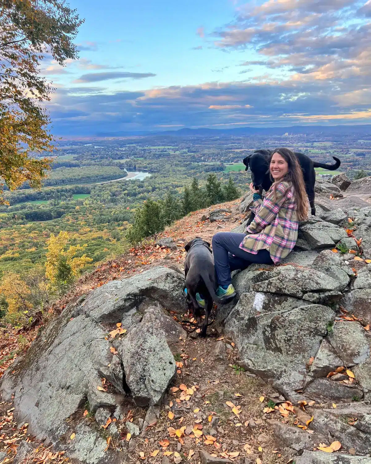 woman in beige flannel and black dog on rocky summit with golden sky in distance in massachusetts.