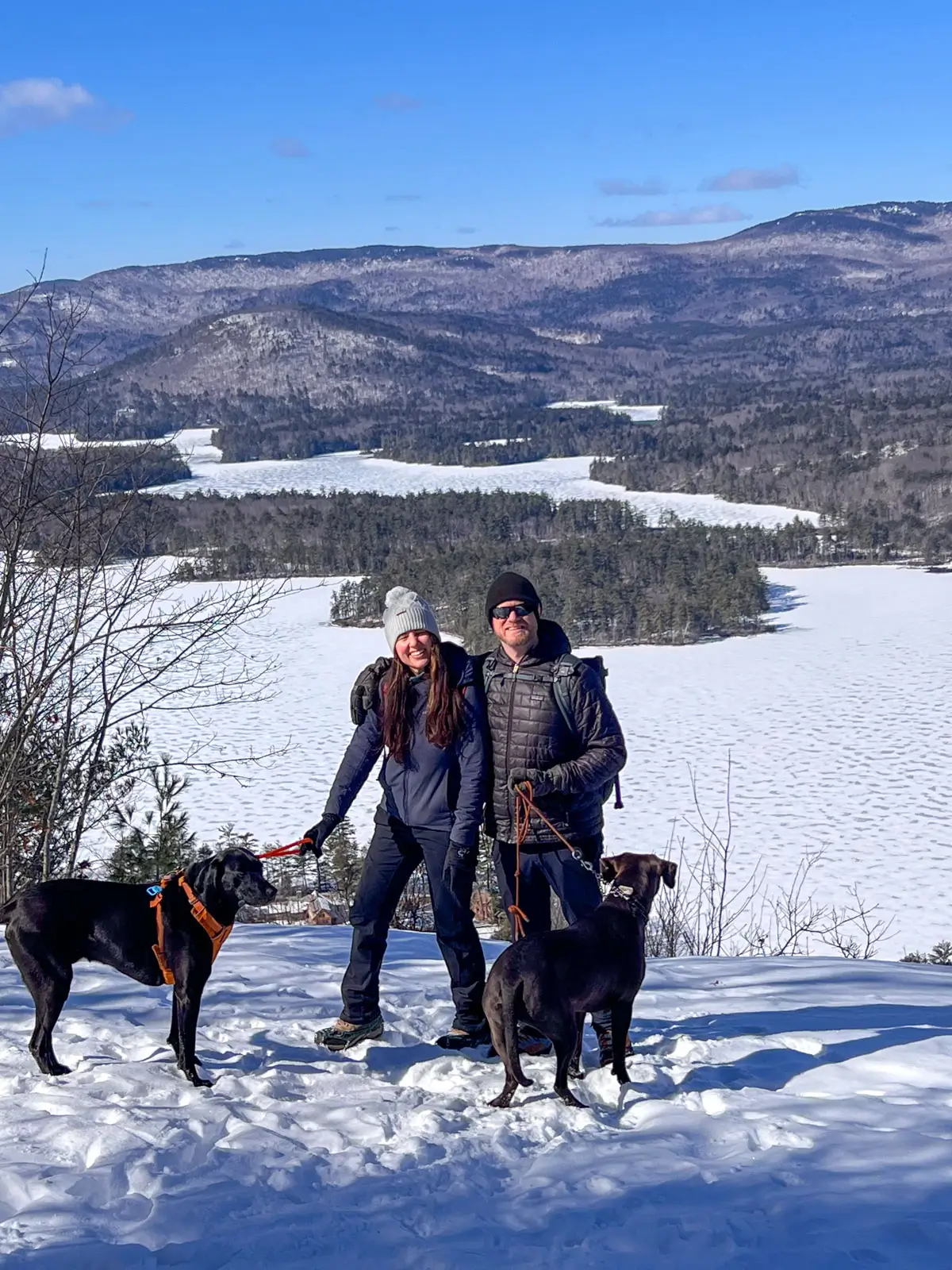 man and woman in snow hats with two big black dogs on snowy summit in new hampshire with frozen white lake in background and brown mountains dusted with snow in distance and blue skies above.