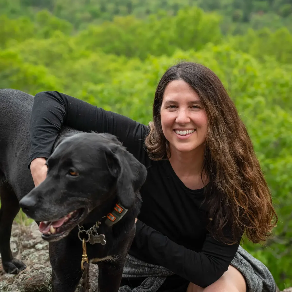 woman with brown hair and in black long sleeve top with arm around black dog on a hike with green trees in the background.