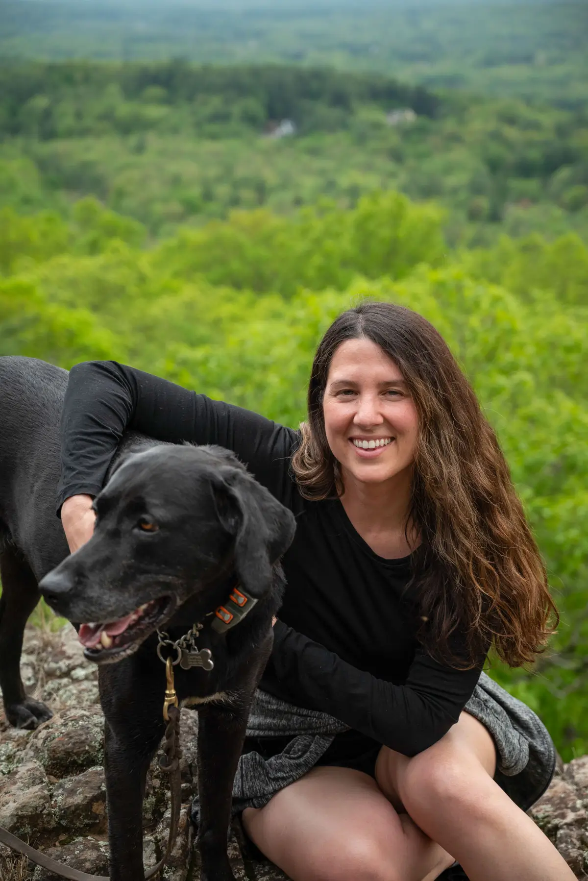 woman with brown hair and in black long sleeve top with arm around black dog on a hike with green trees in the background.
