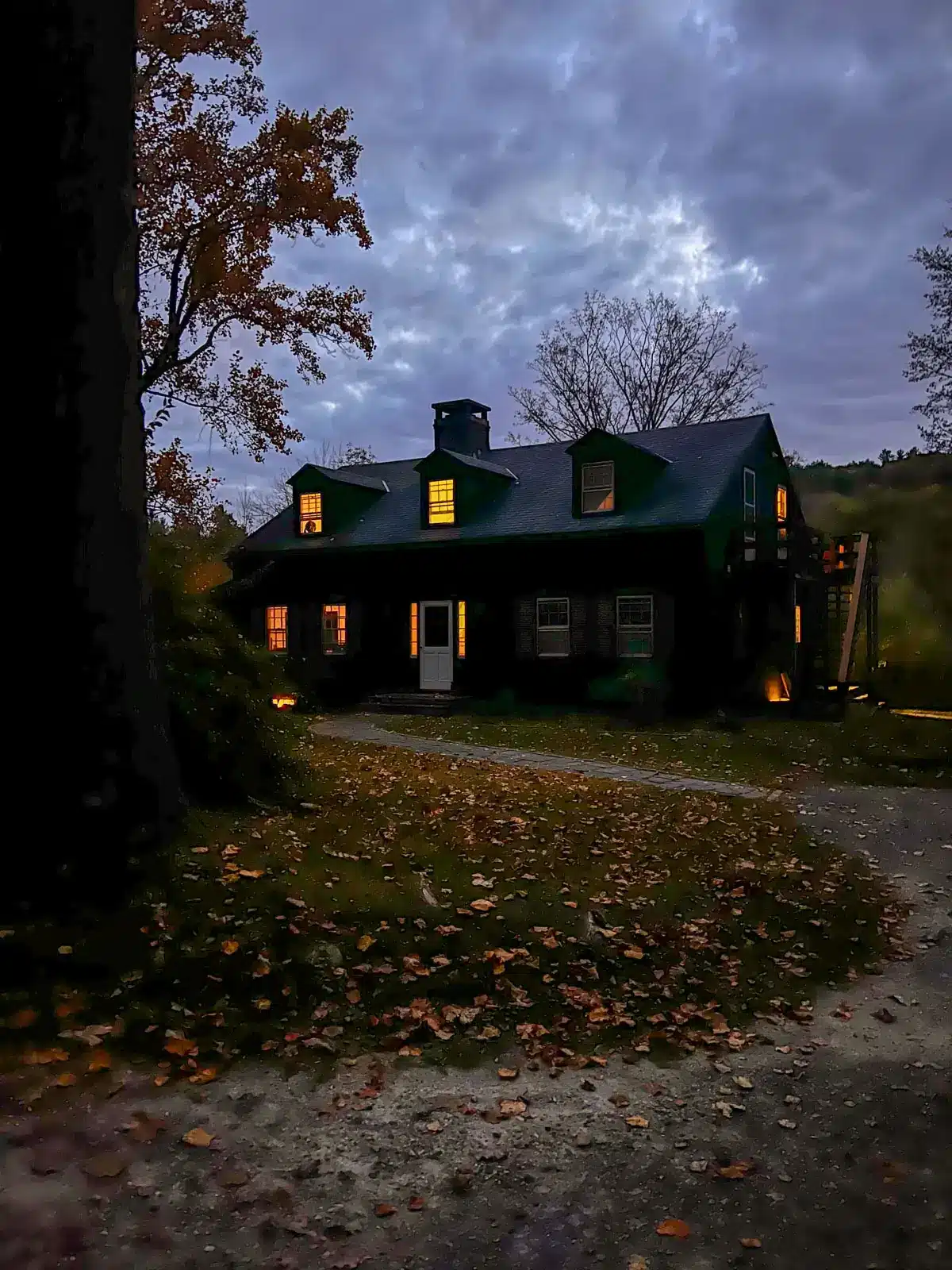 house at dusk with golden lights shining from within with a spooky vibe and orange leaves all around.
