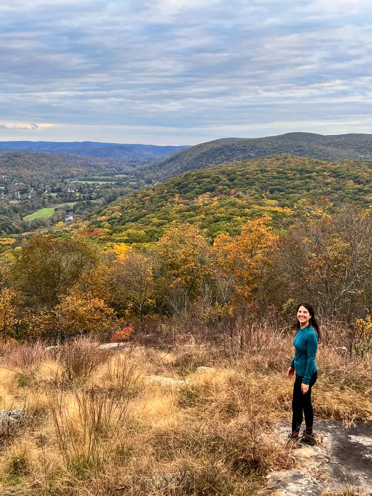 woman in long sleeved blue shirt at the top of a hike in kent at caleb's peak with orange colroed leaves in the distance.